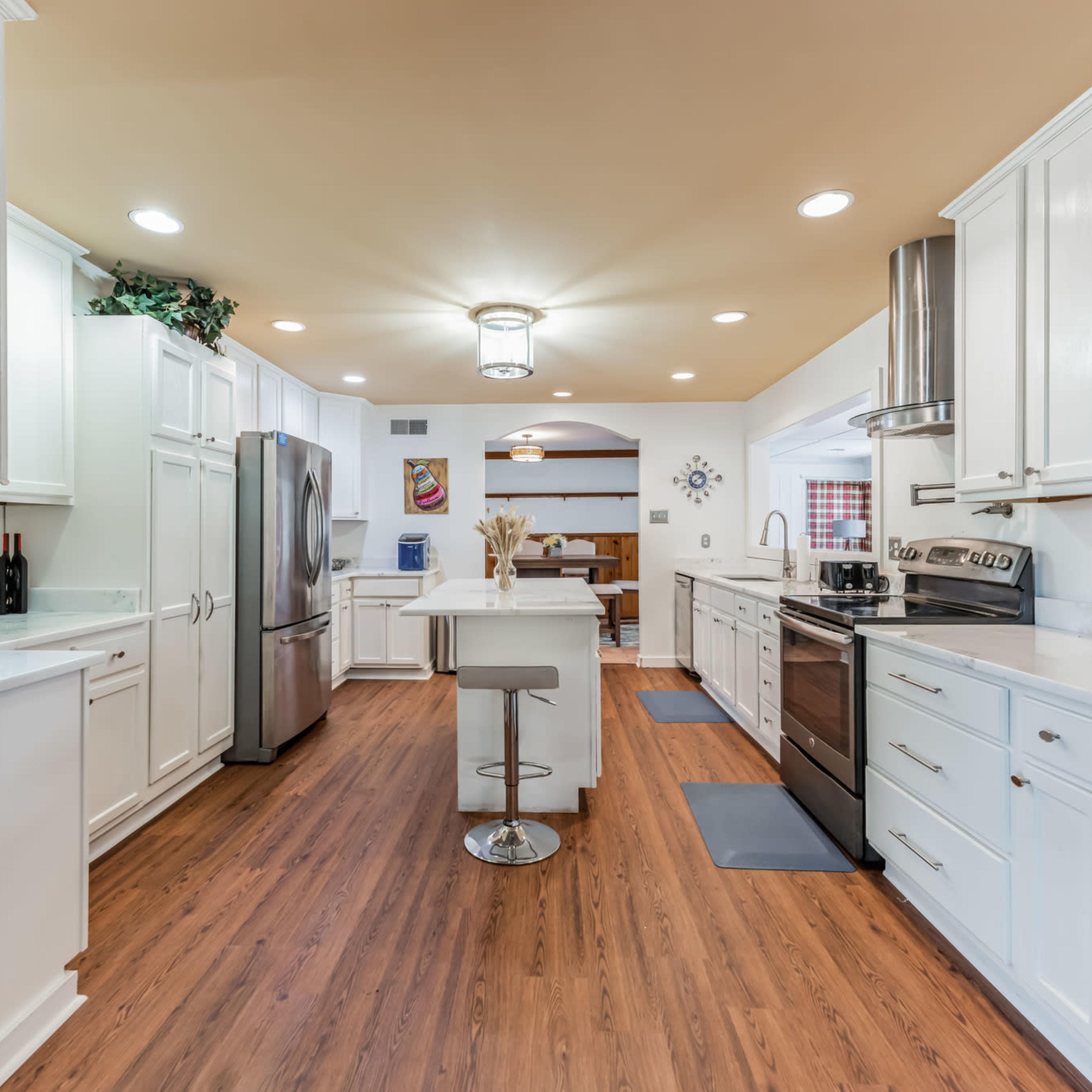 A modern kitchen featuring white cabinetry, stainless steel appliances, and a central island with a stool, all set against a wood-paneled wall and warm-toned flooring.