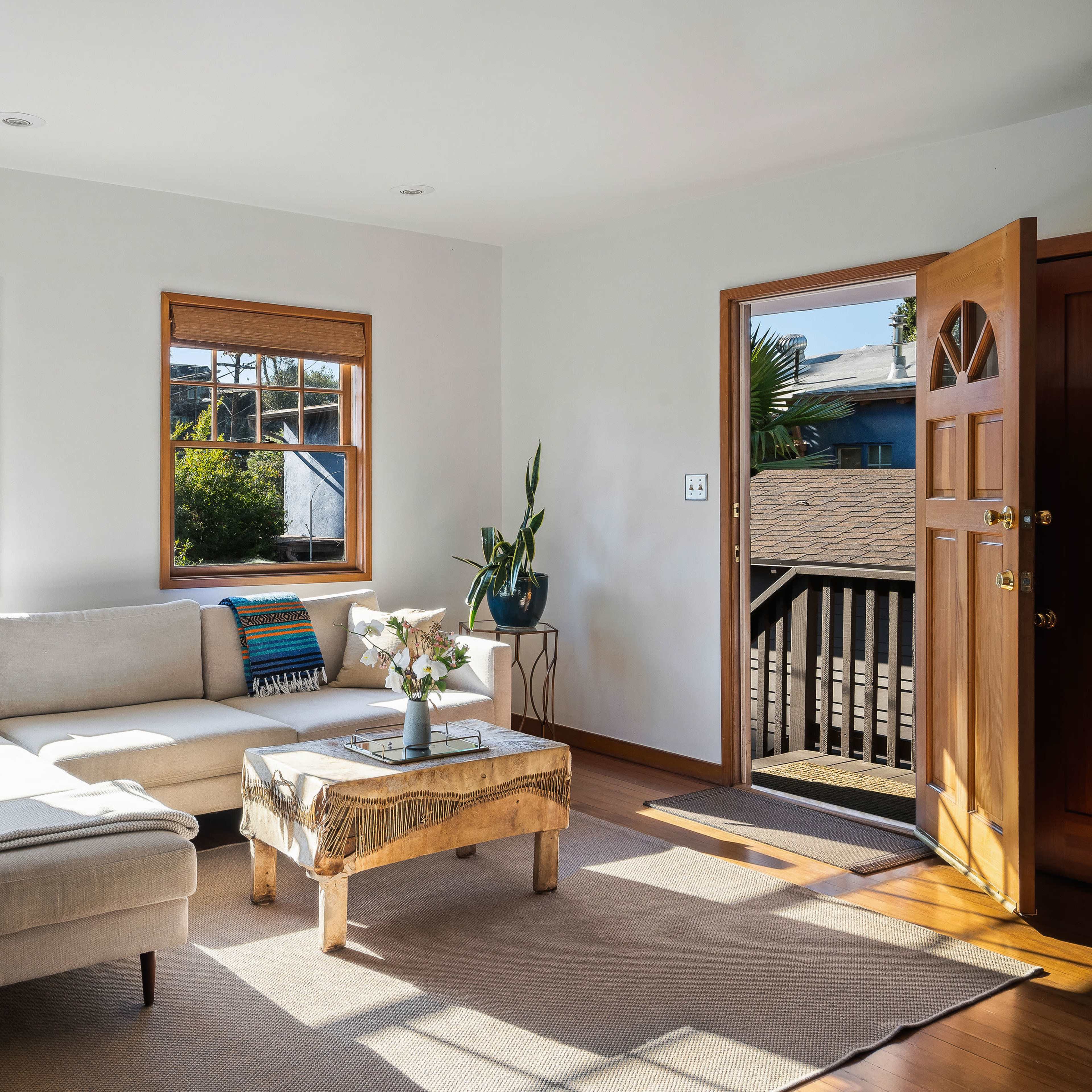 A light-filled living room features a sectional sofa, a coffee table, and three windows with views of the outside.