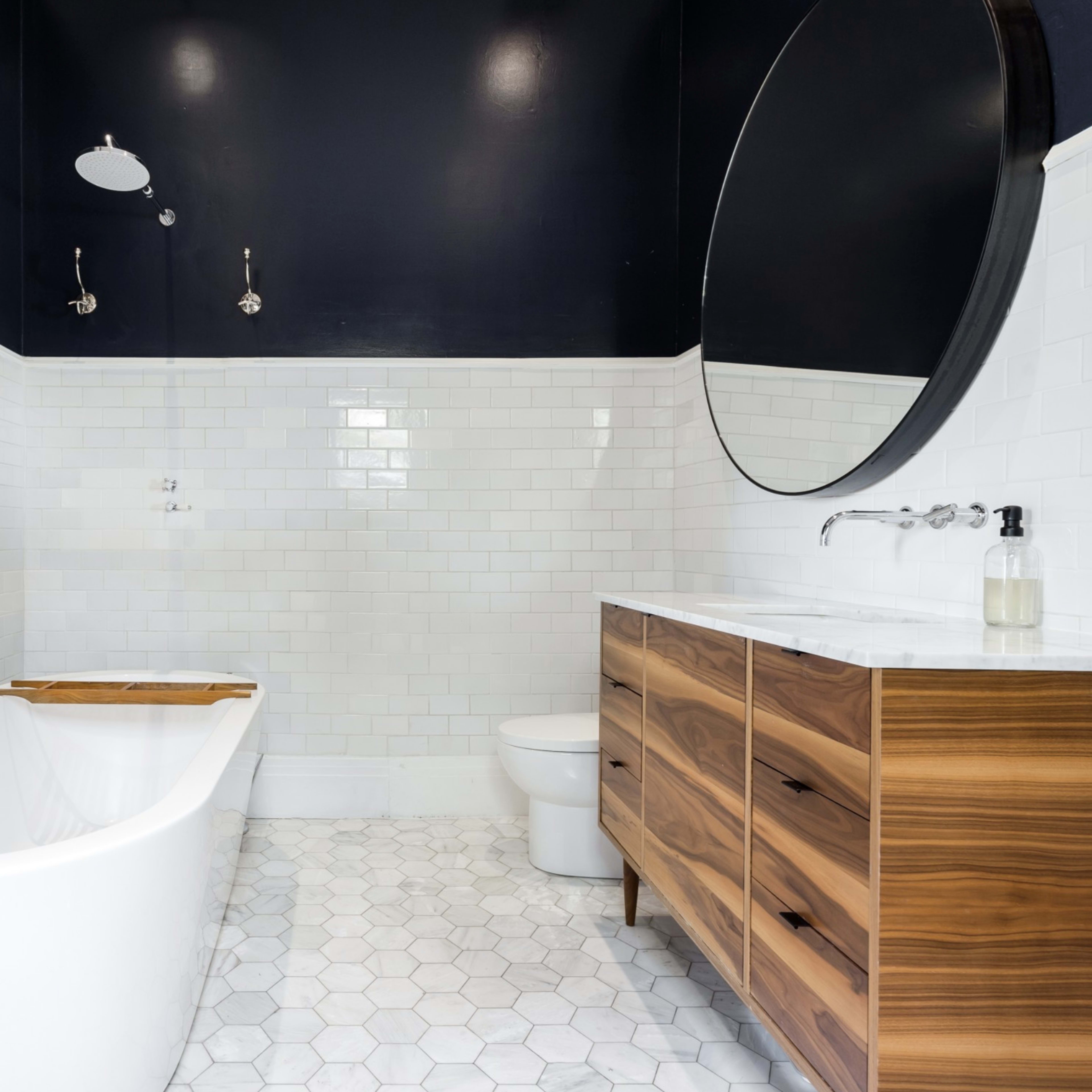 A modern bathroom featuring a freestanding bathtub, a circular mirror, a wooden vanity with multiple drawers, and white tile walls.