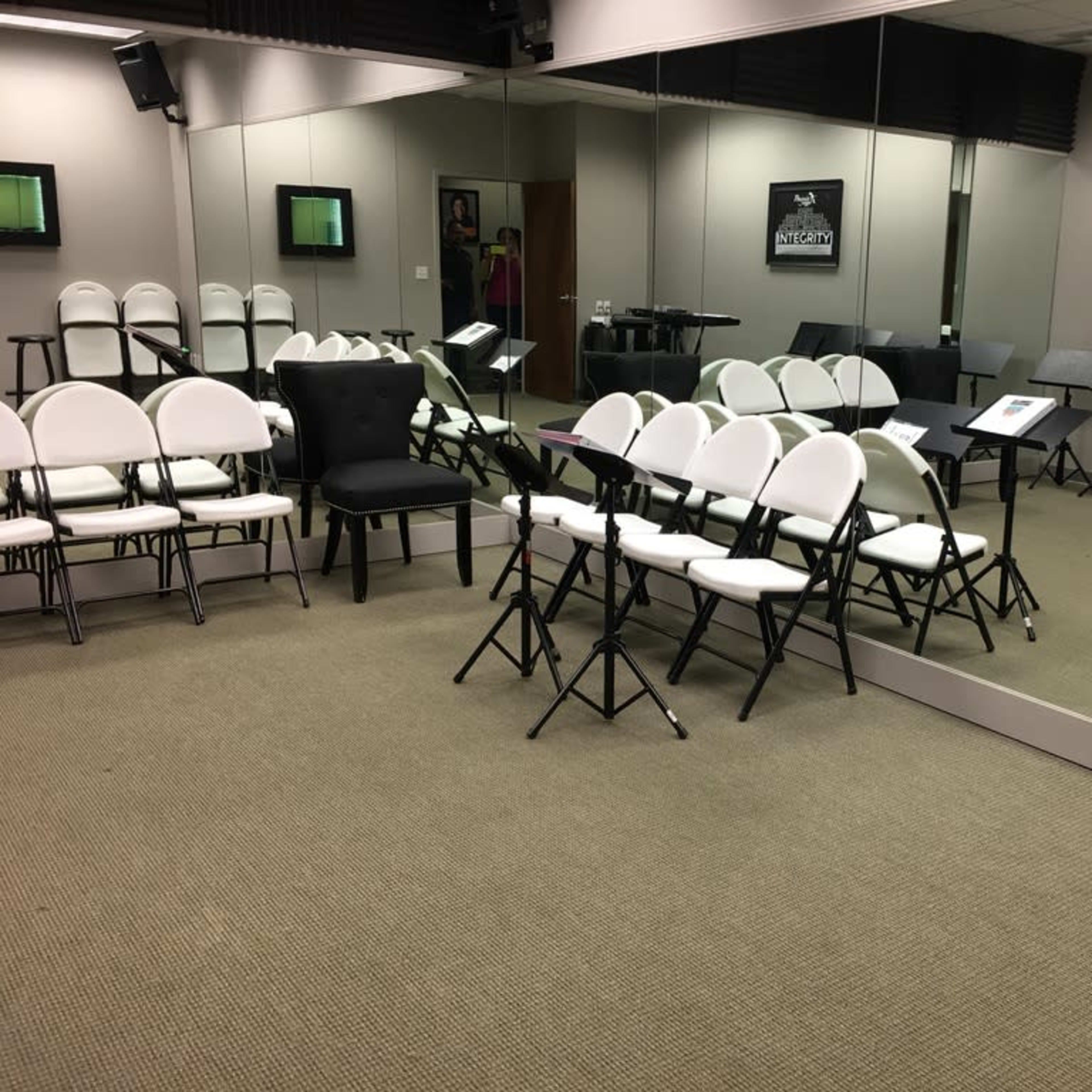 A music rehearsal room features several rows of white and black chairs, music stands, and large mirrors on the walls.