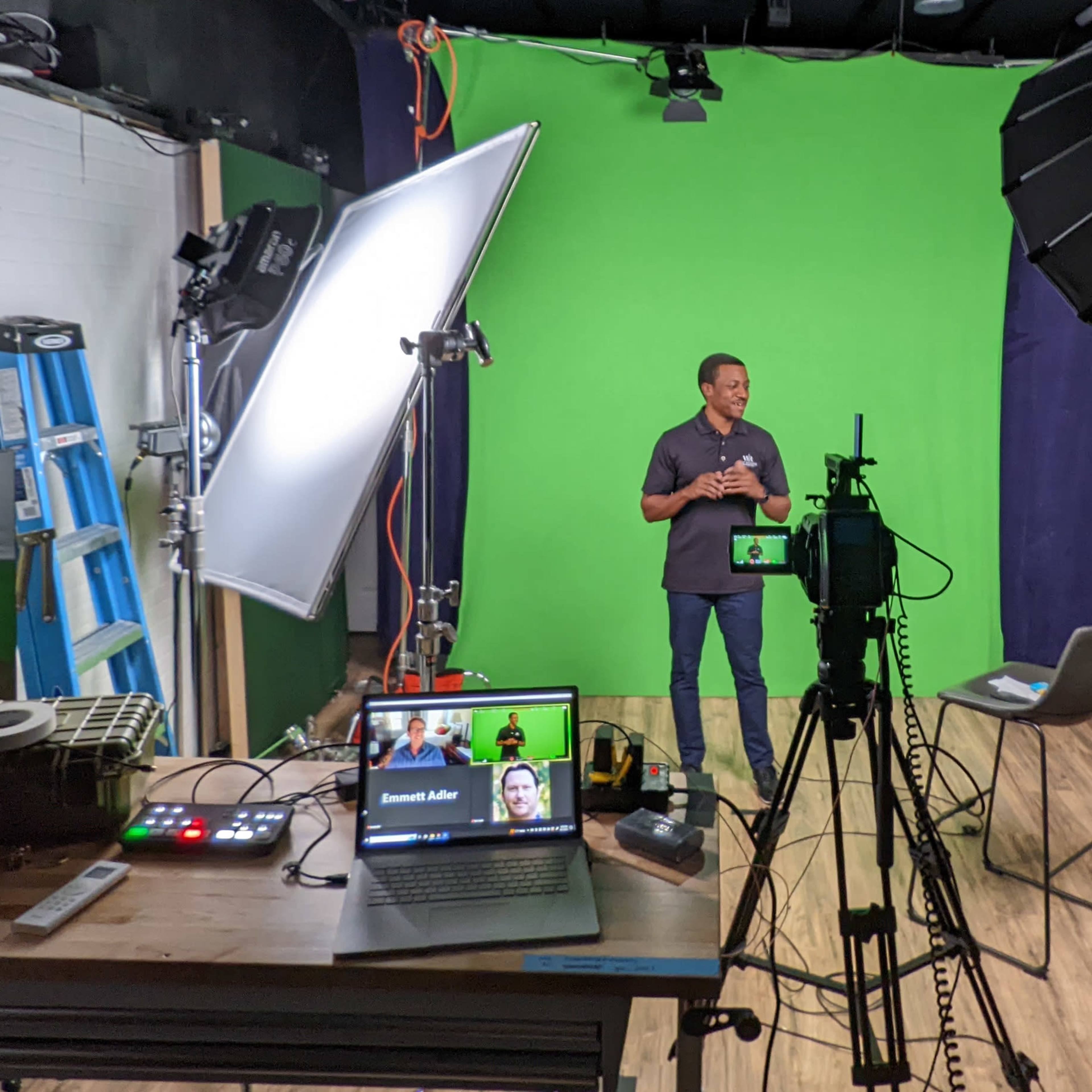 A person stands in front of a green backdrop in a studio, with lighting equipment and a laptop displaying a video call setup on a table.