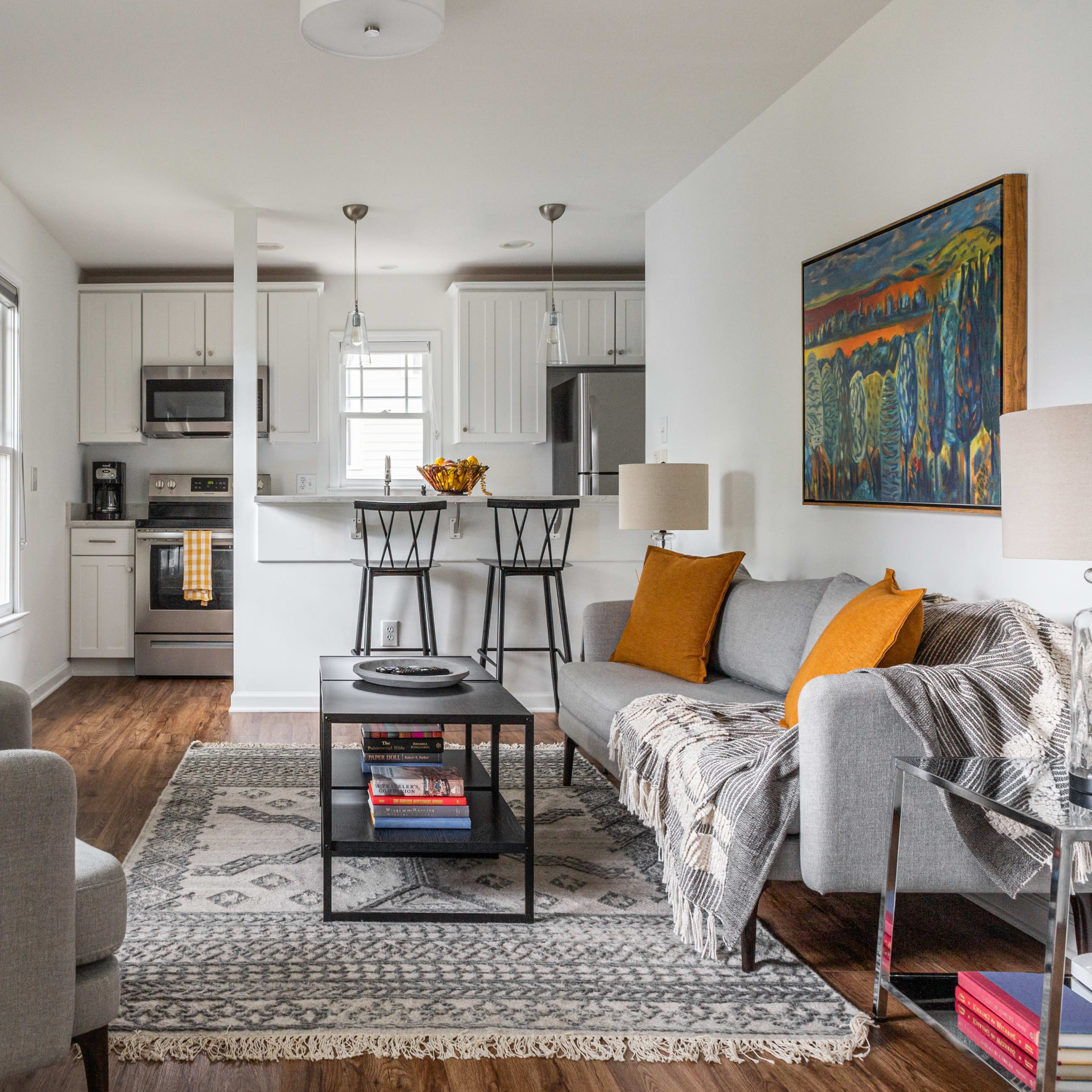 A bright living area featuring a gray sofa, an accent chair, a coffee table with books, and a kitchenette in the background.