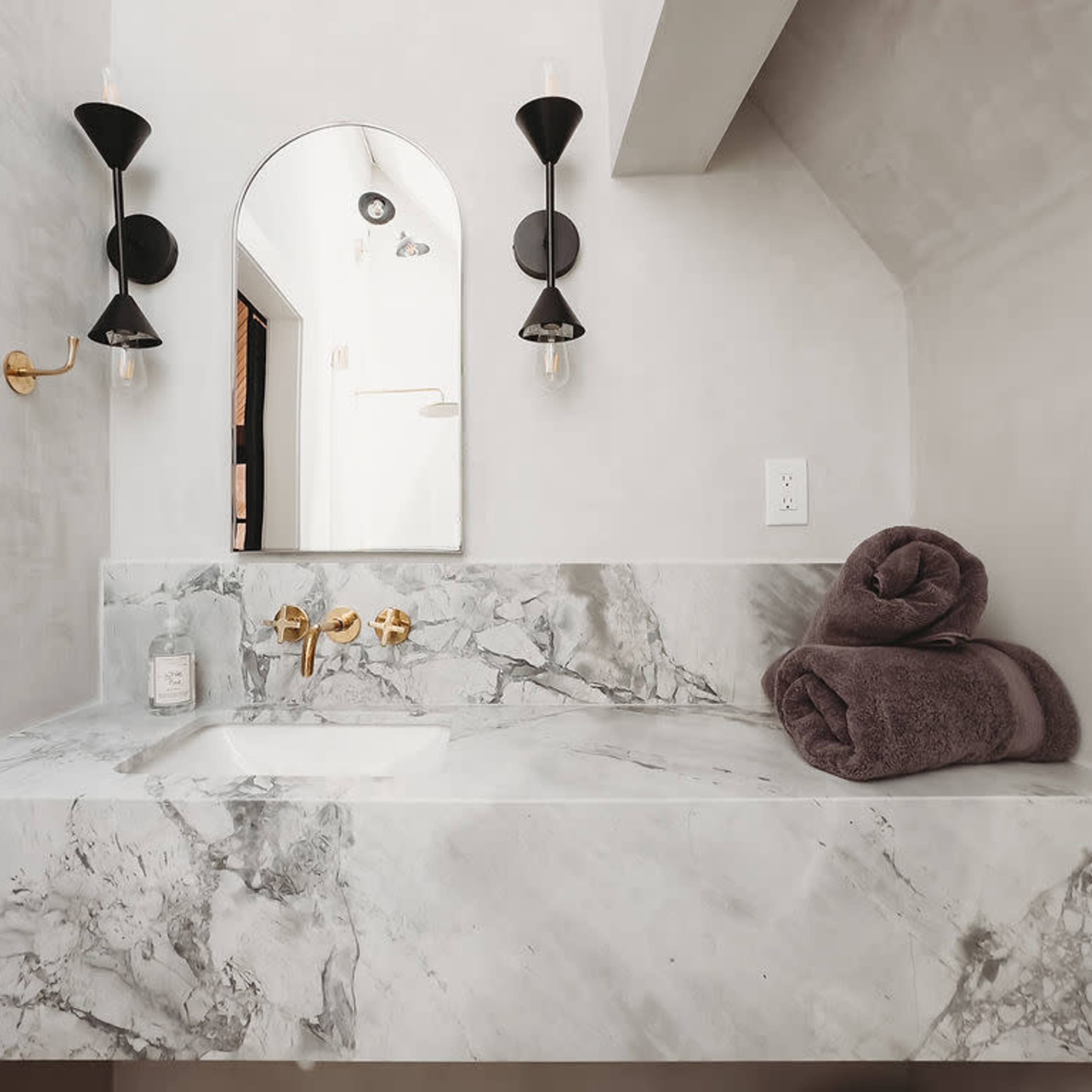 A minimalist bathroom featuring a marble countertop, a large mirror, gold fixtures, and neatly rolled towels.