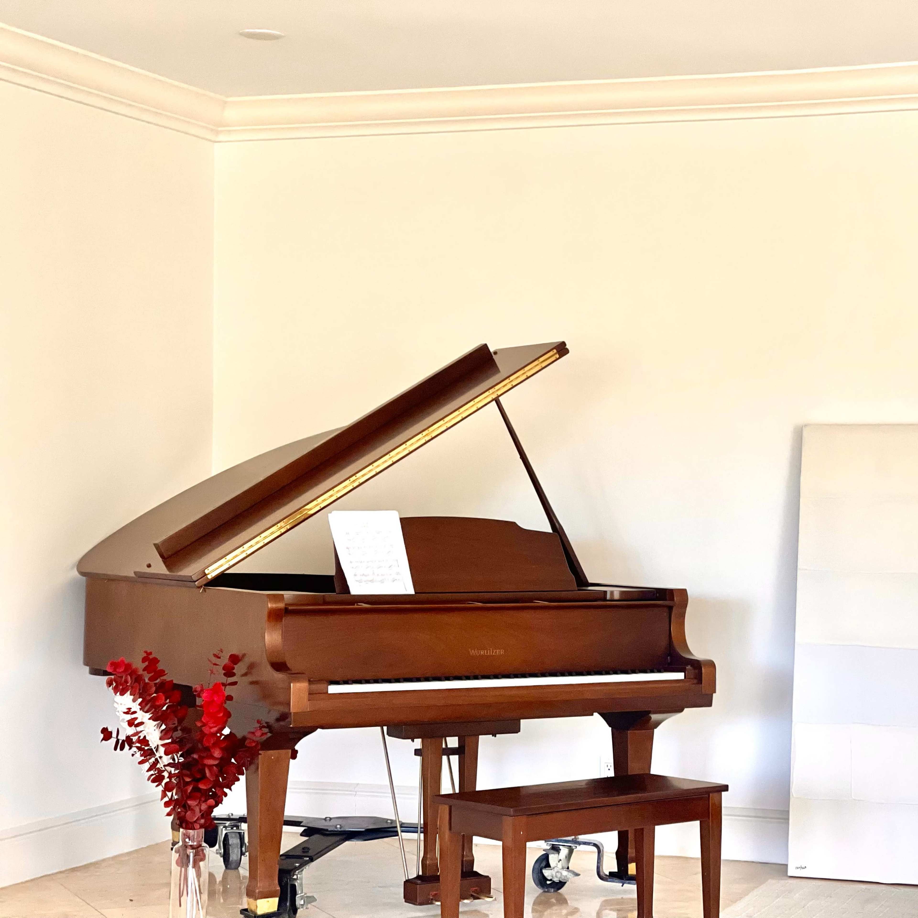 A grand piano with its lid open sits next to a white canvas and a vase of red flowers in a light-filled room.