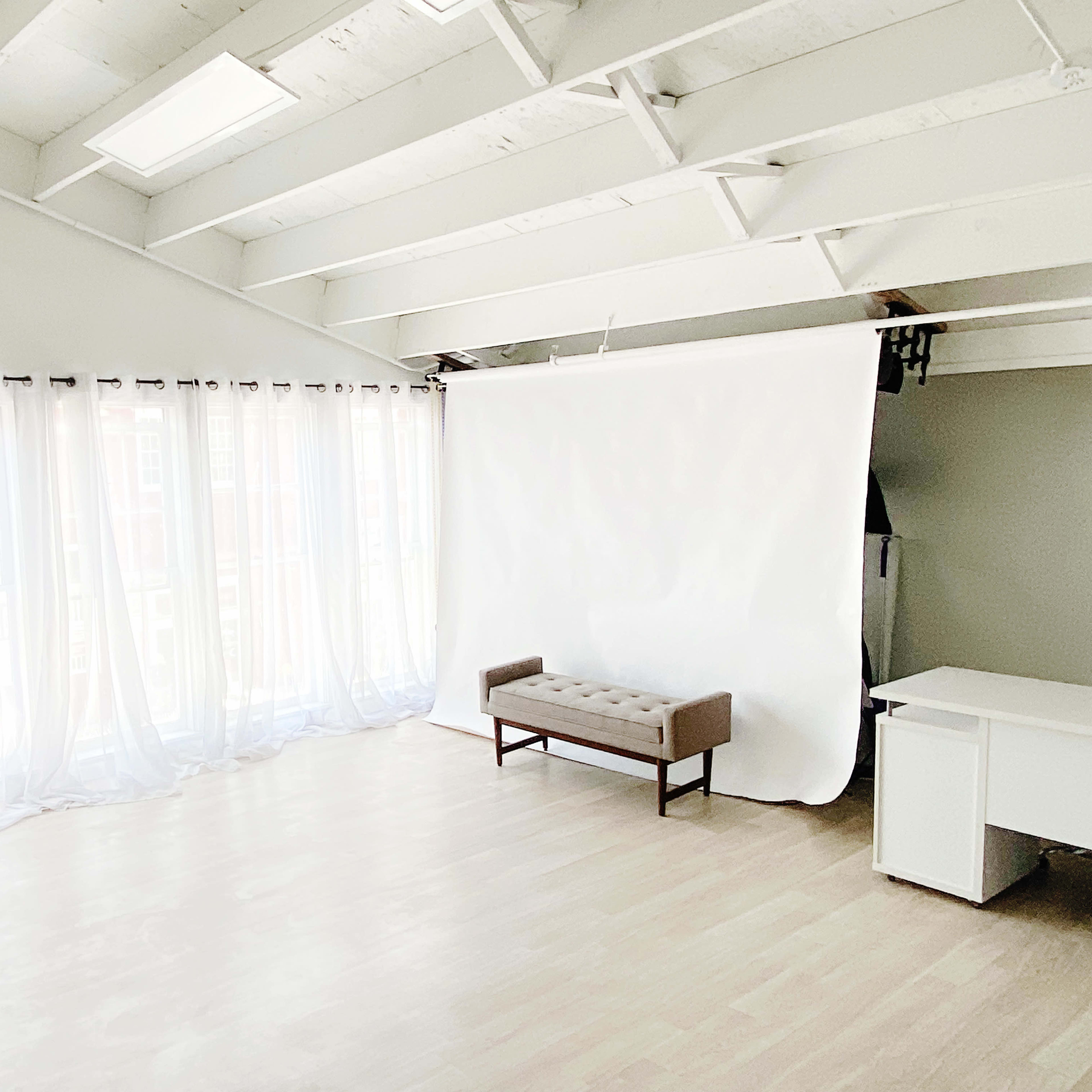 A bright, spacious room features large windows with sheer white curtains, a light-colored wooden floor, a simple beige bench, and a white backdrop against one wall.