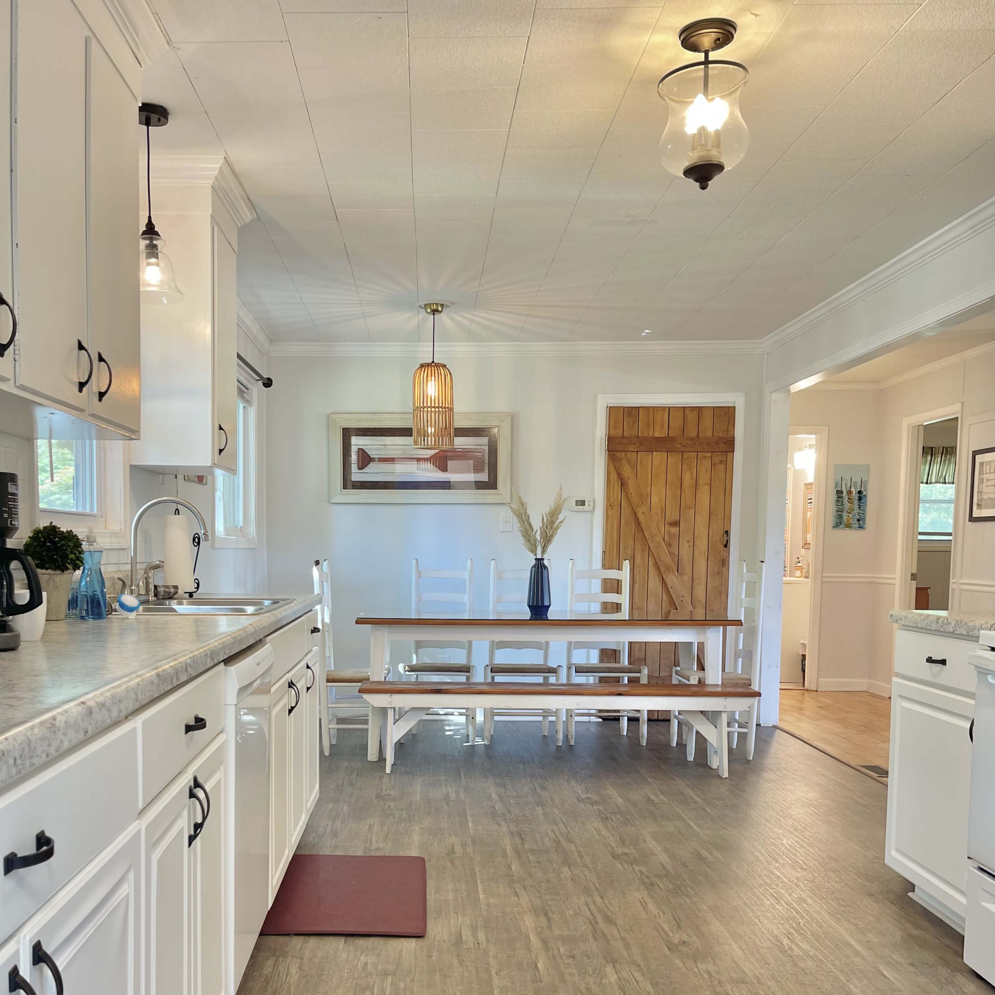 A bright, open kitchen and dining area featuring white cabinetry, a large wooden table with benches, and a door leading to another room.
