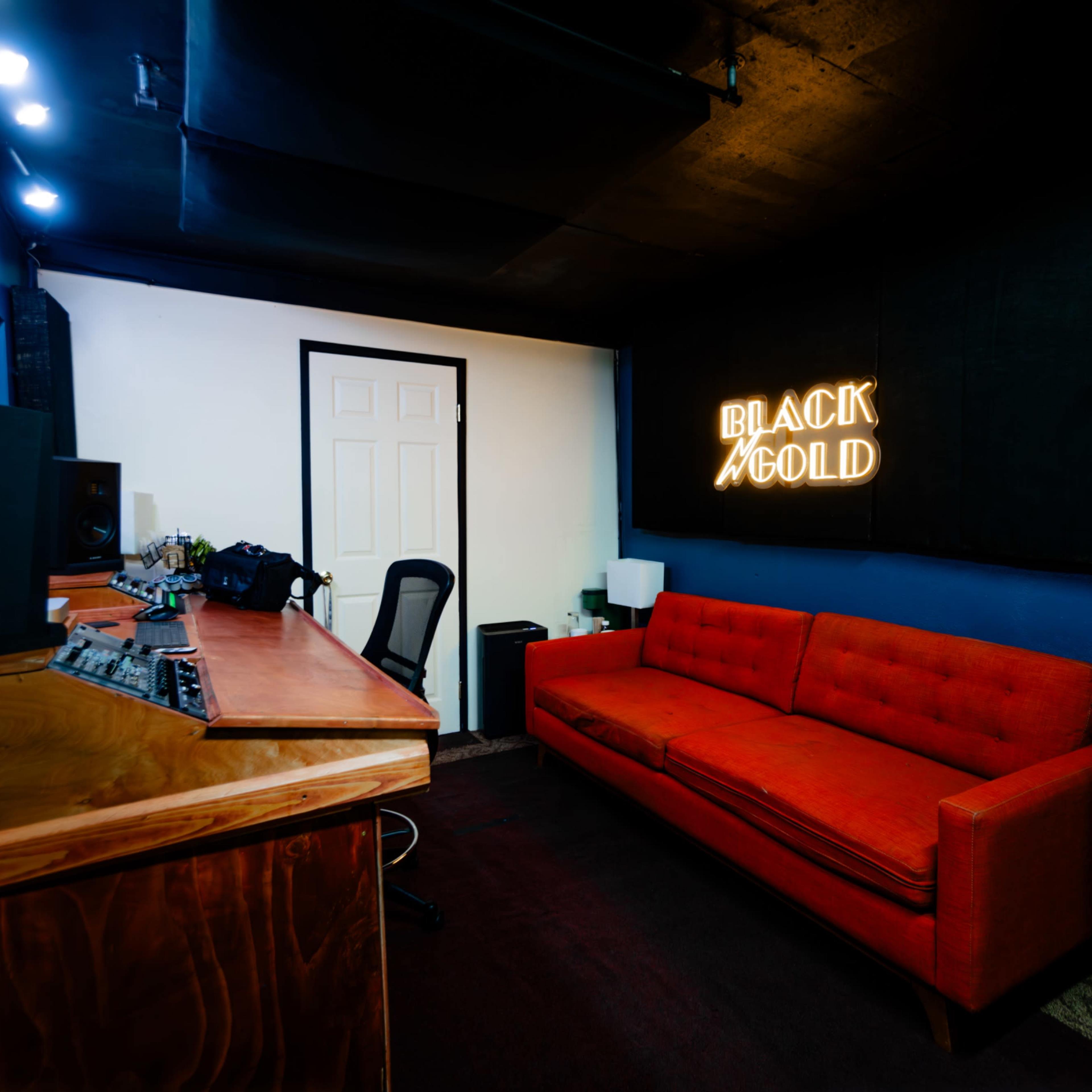 A music studio featuring a red sofa, a wooden desk with audio equipment, and a neon sign that reads "BLACK GOLD."