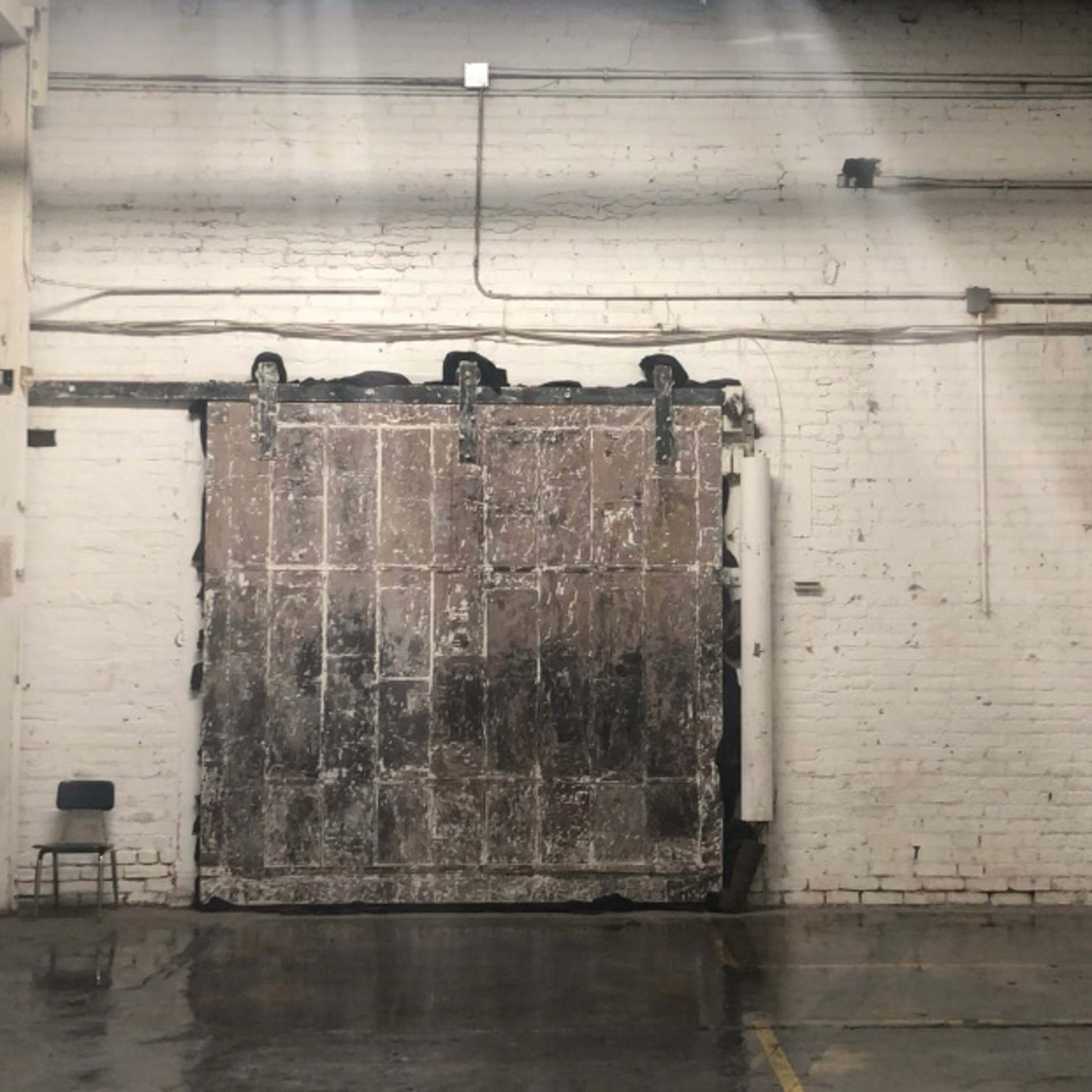 A large, weathered sliding door in an industrial space with exposed brick walls and a polished concrete floor, accompanied by a few empty chairs in the foreground.