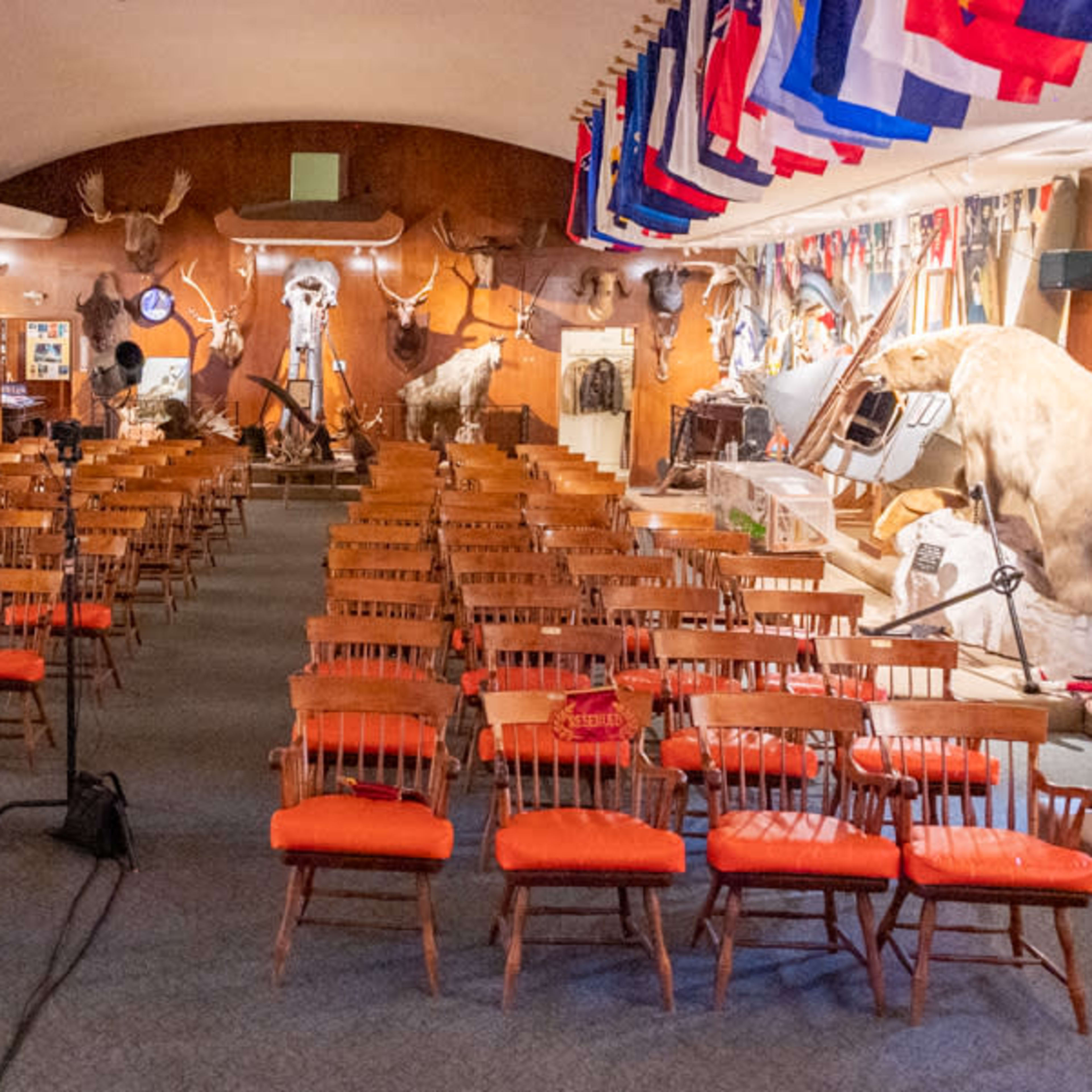 A large room filled with rows of wooden chairs, decorated with mounted animal trophies and flags hanging from the ceiling.