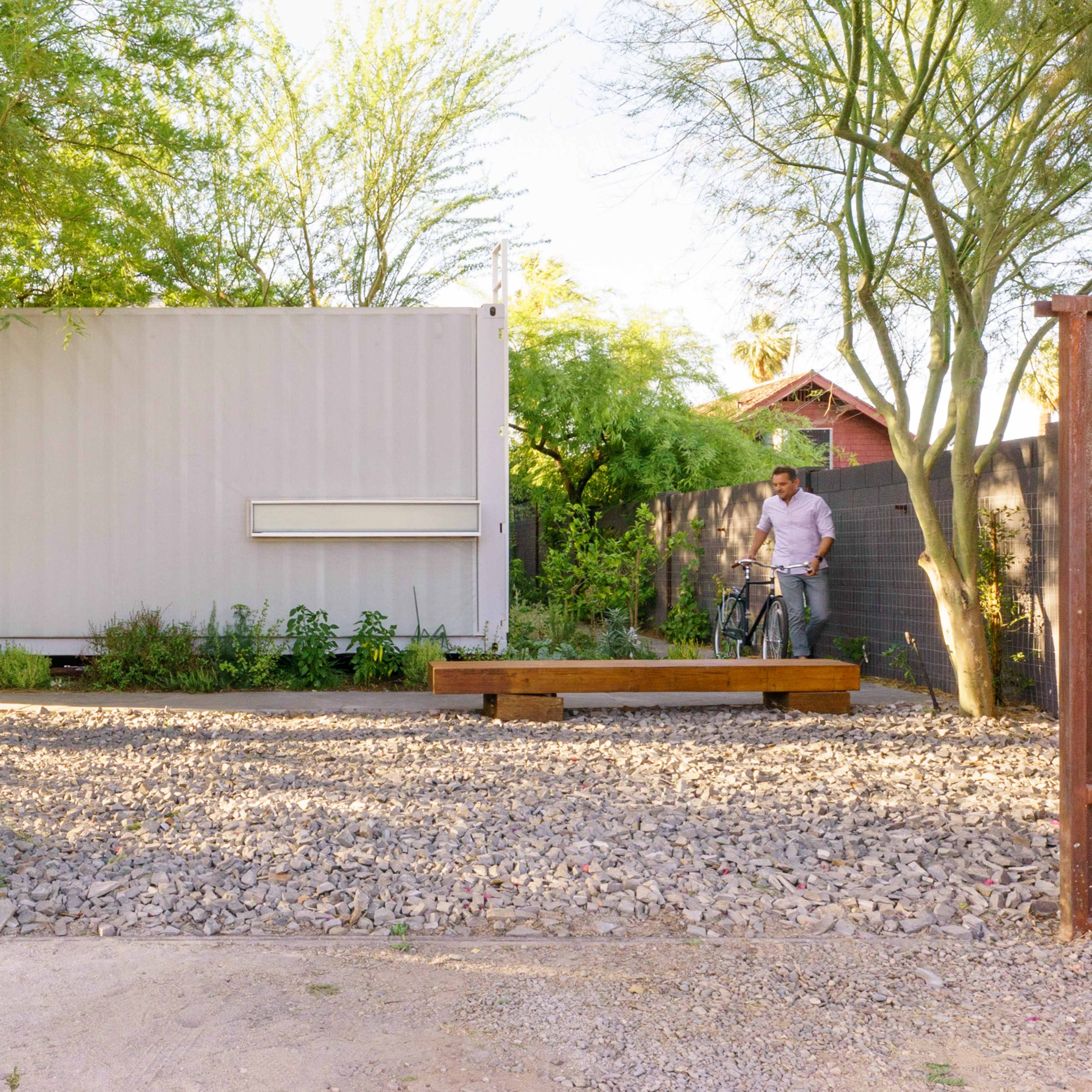 A person standing next to a bicycle in a gravel courtyard surrounded by trees and a modern building.