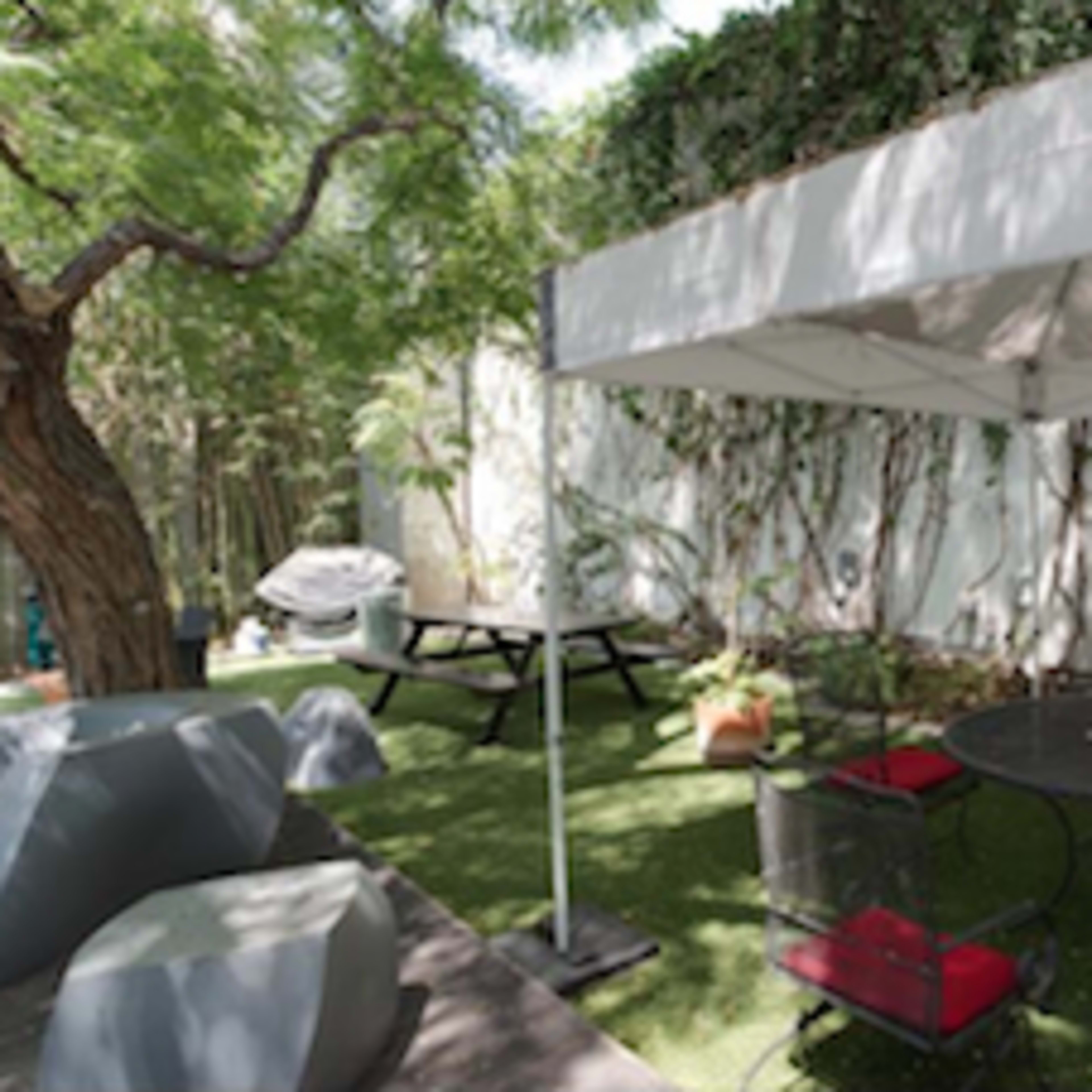 A shaded outdoor seating area features a round table with chairs, surrounded by greenery and a barbecue grill in the background.