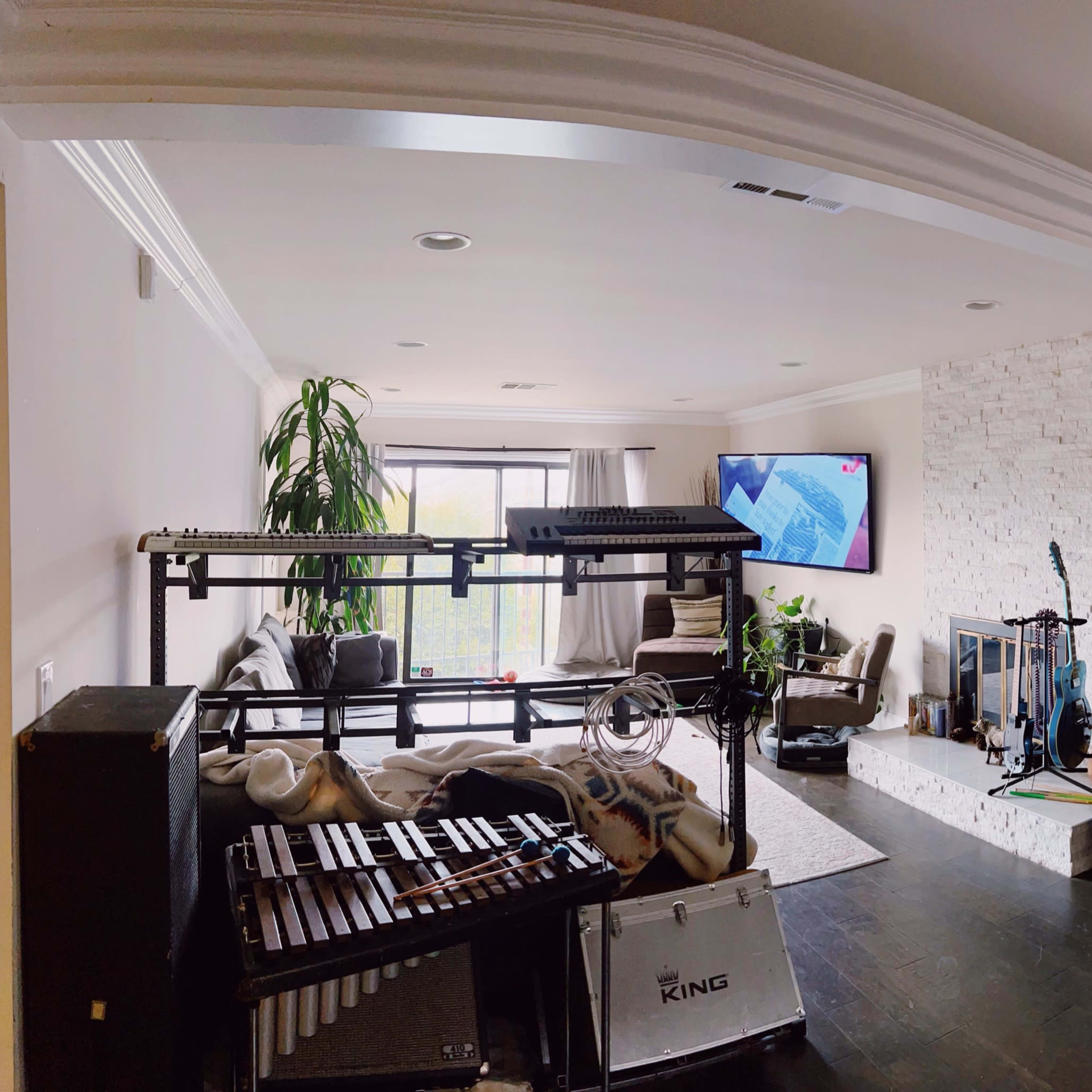 A spacious music studio featuring various instruments, including keyboards and a xylophone, with a large window and modern furniture in the background.