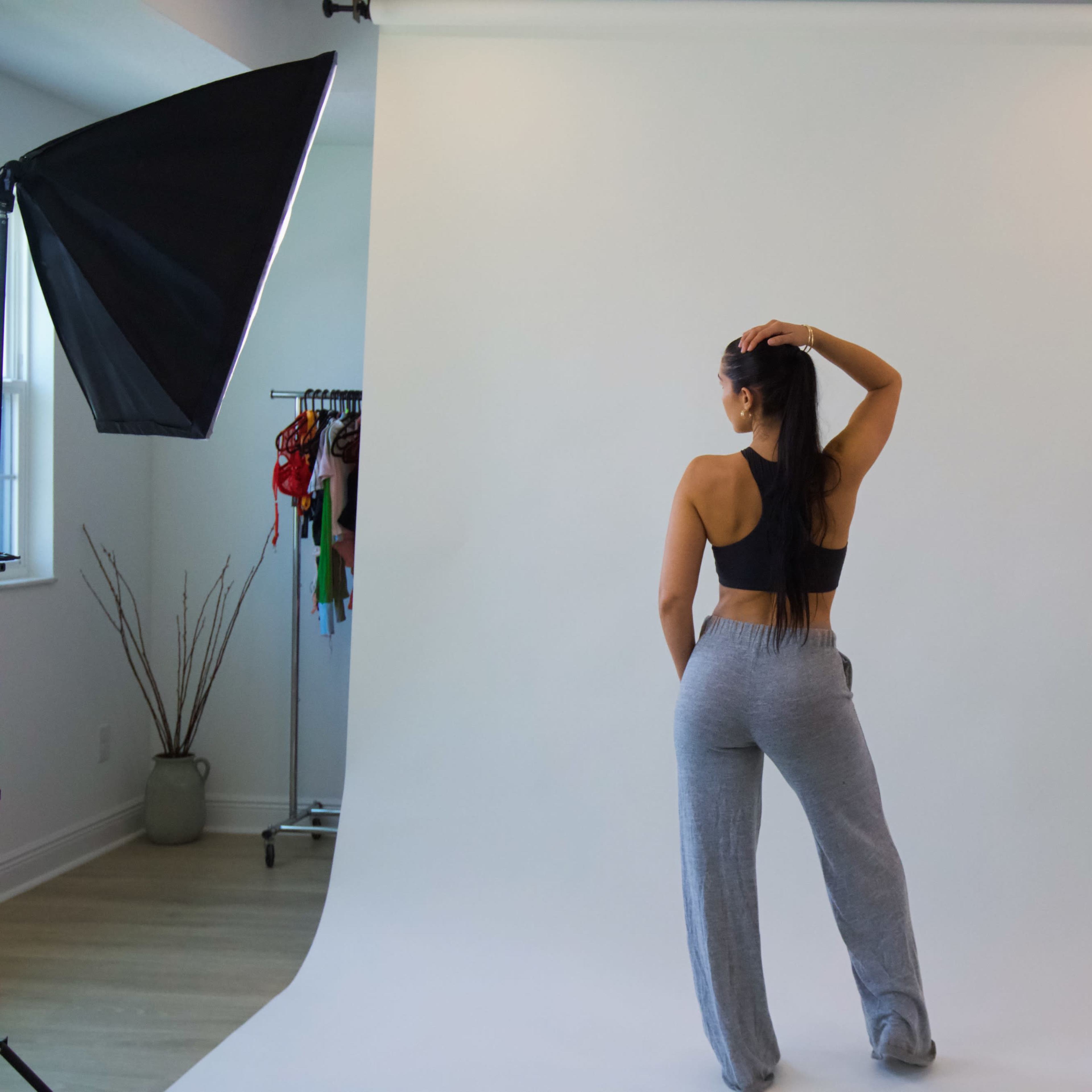 A person is posing with their back to the camera in a studio setting, with softbox lights on either side and a plain backdrop.