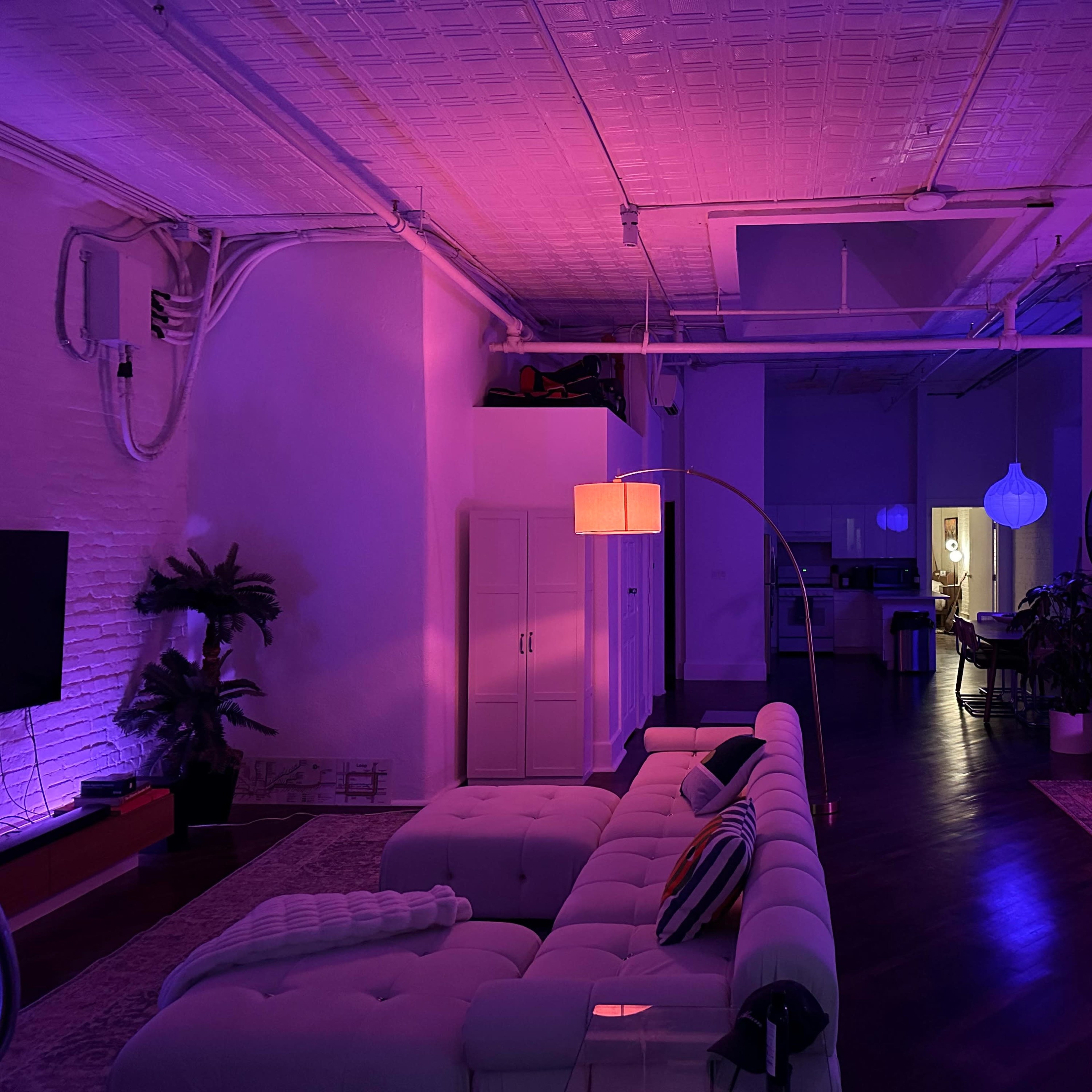 A spacious room is illuminated with purple lighting, featuring a white couch, a television, and a doorway leading to a kitchen area.