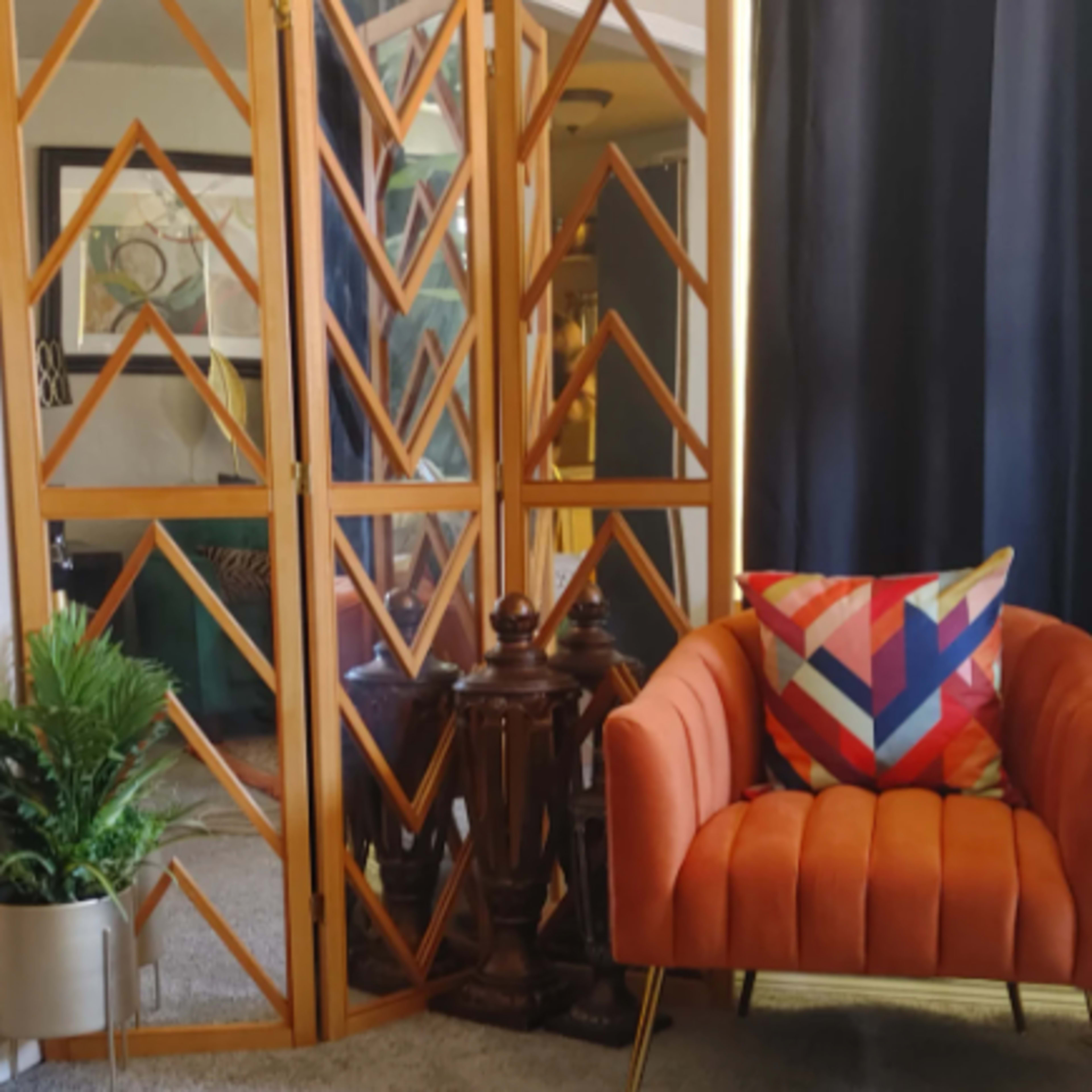 A modern living space features an orange tufted sofa, a geometric pillow, a decorative screen, and a potted plant.