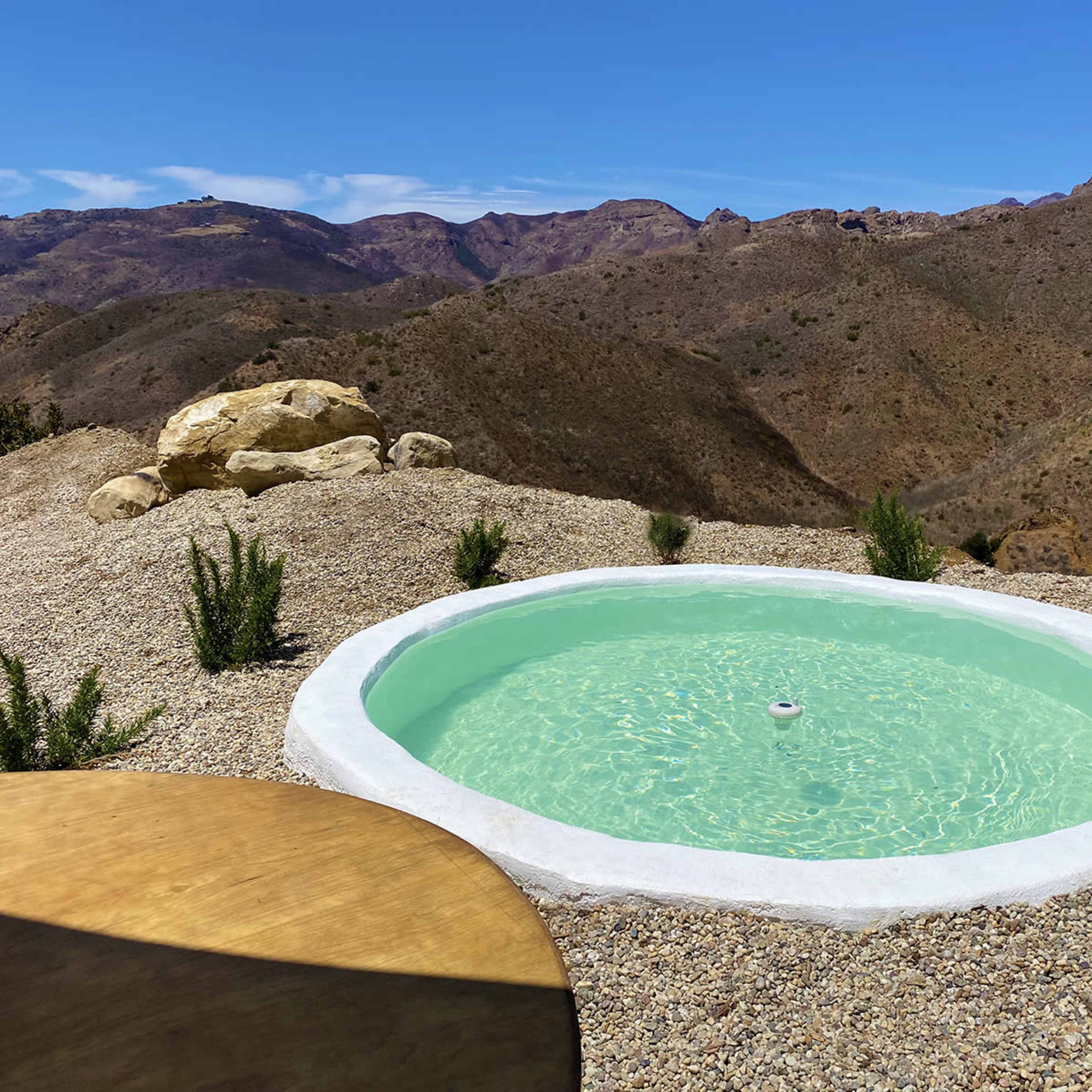 A circular outdoor pool filled with light green water is situated on a rocky hillside overlooking mountainous terrain.