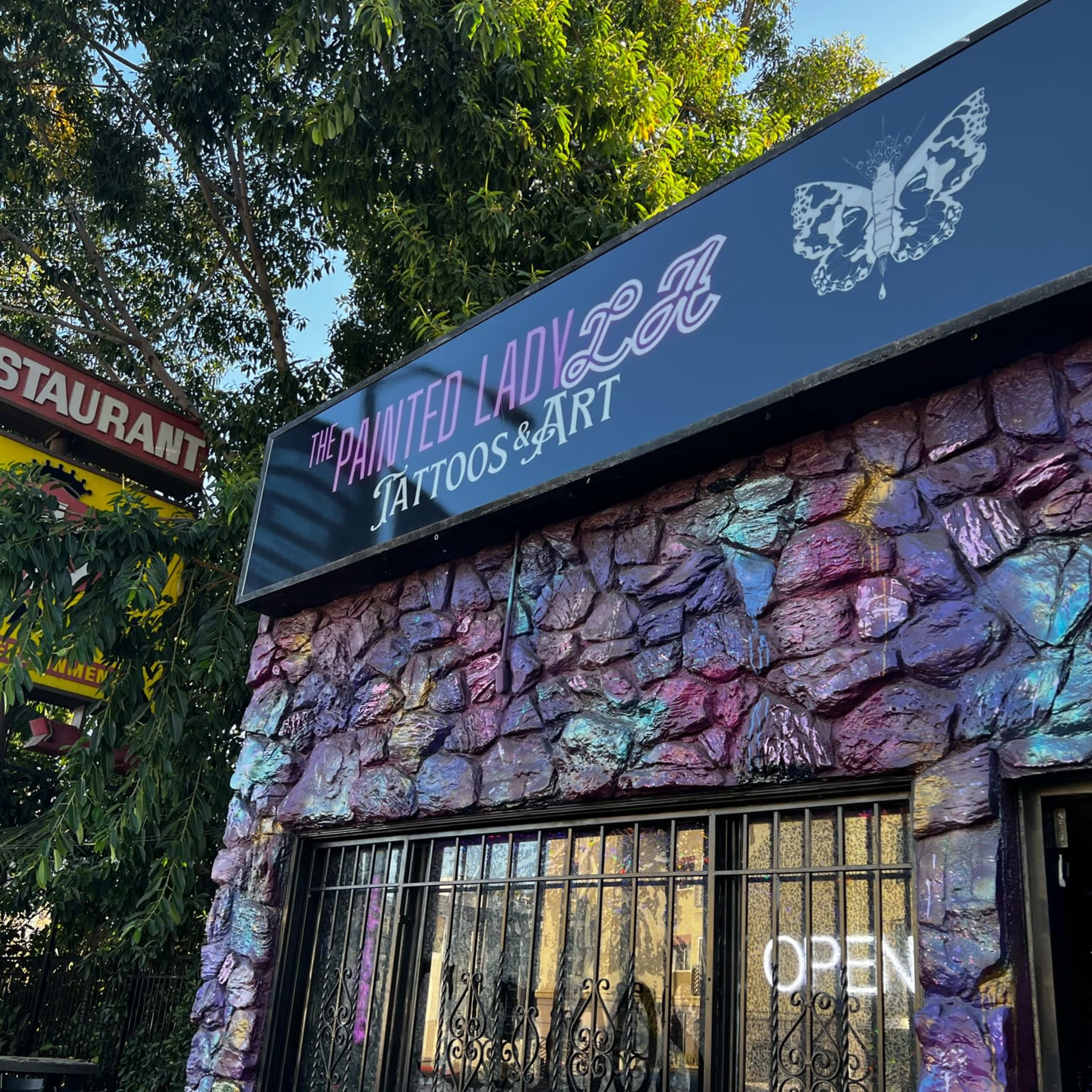 A tattoo shop called "The Painted Lady LA," featuring a colorful stone exterior and a sign with a butterfly motif.