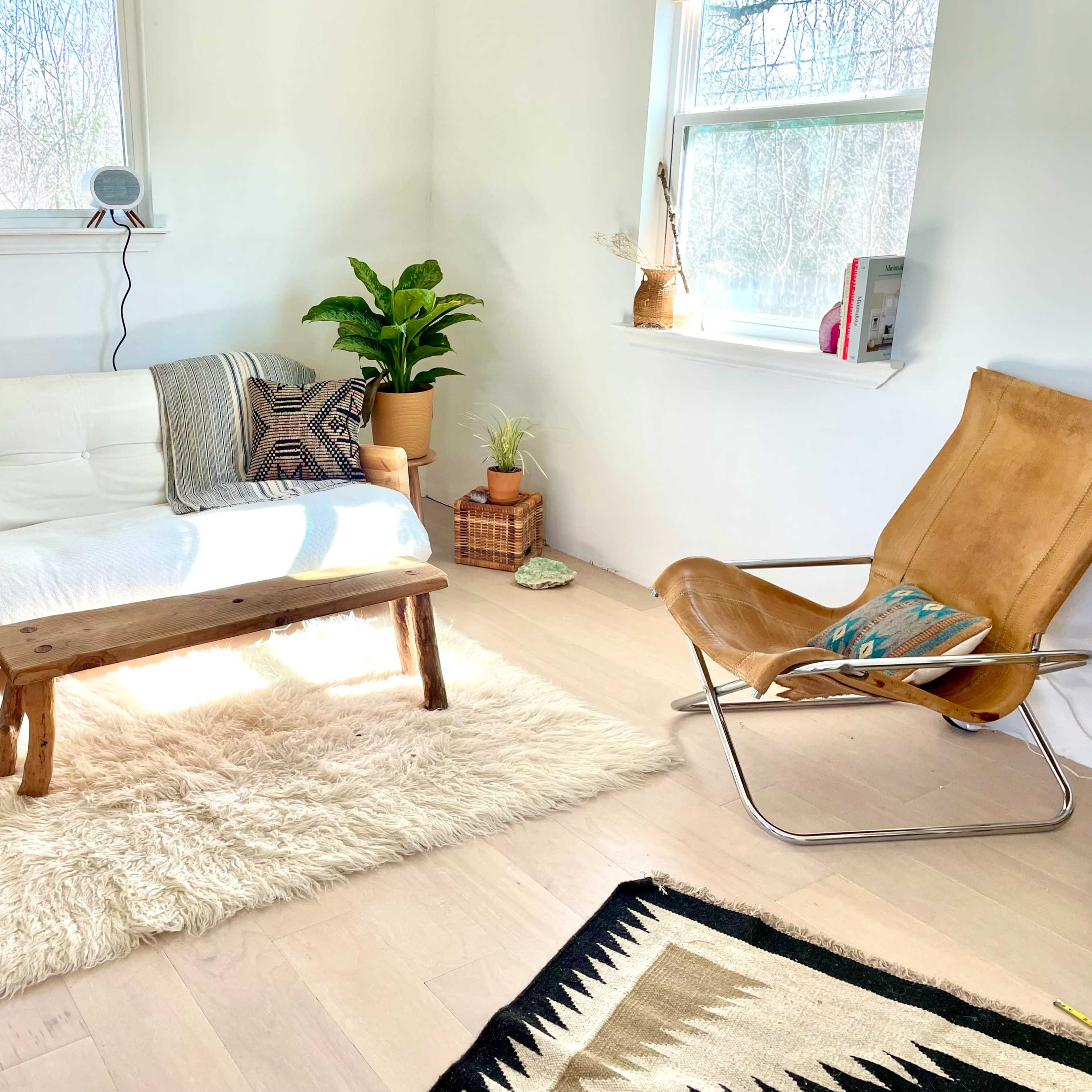 A bright, minimalist living room featuring a white sofa, a wooden coffee table, a beige rug, and a leather chair.