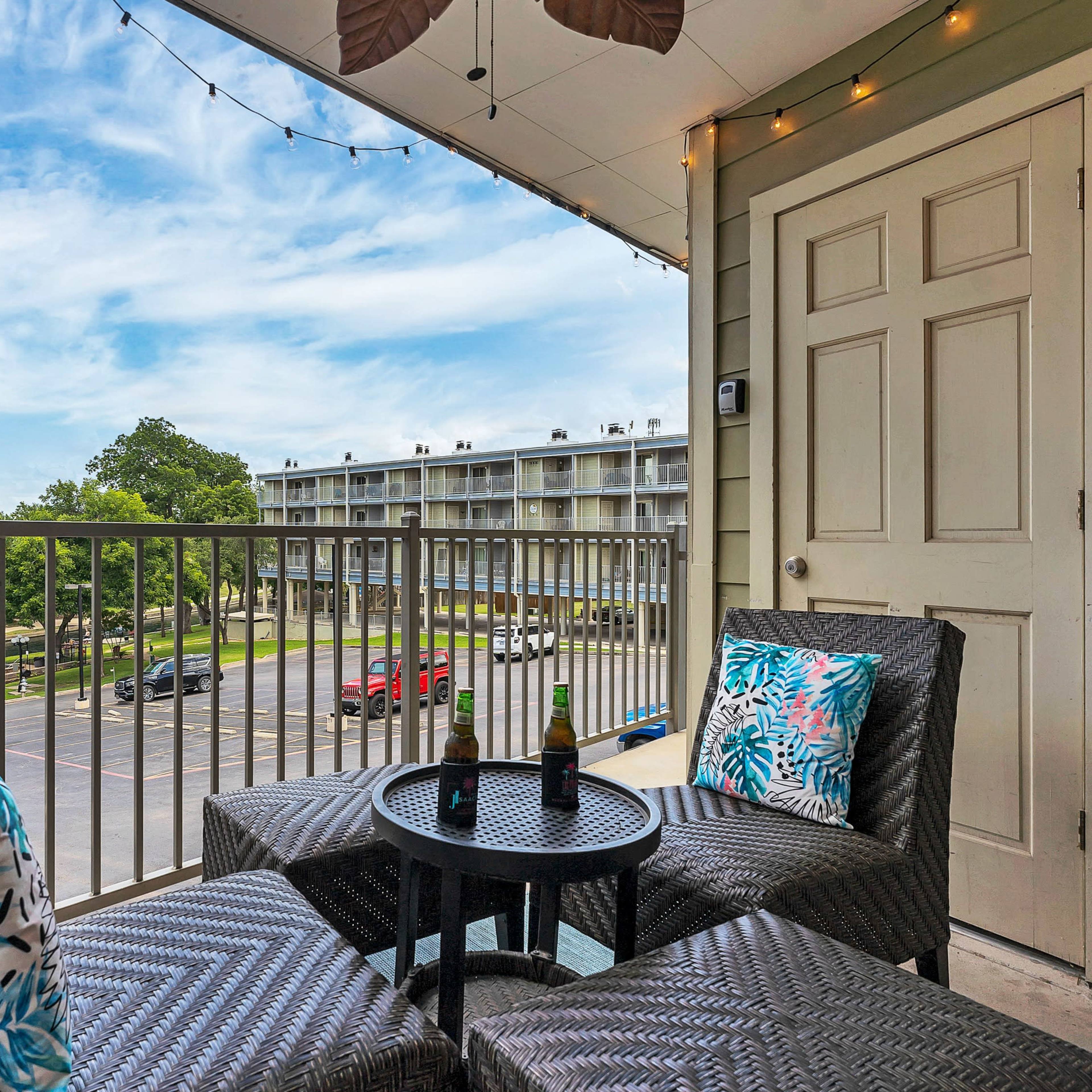 A balcony with two wicker chairs and a small table overlooks a parking area and a building in the distance.