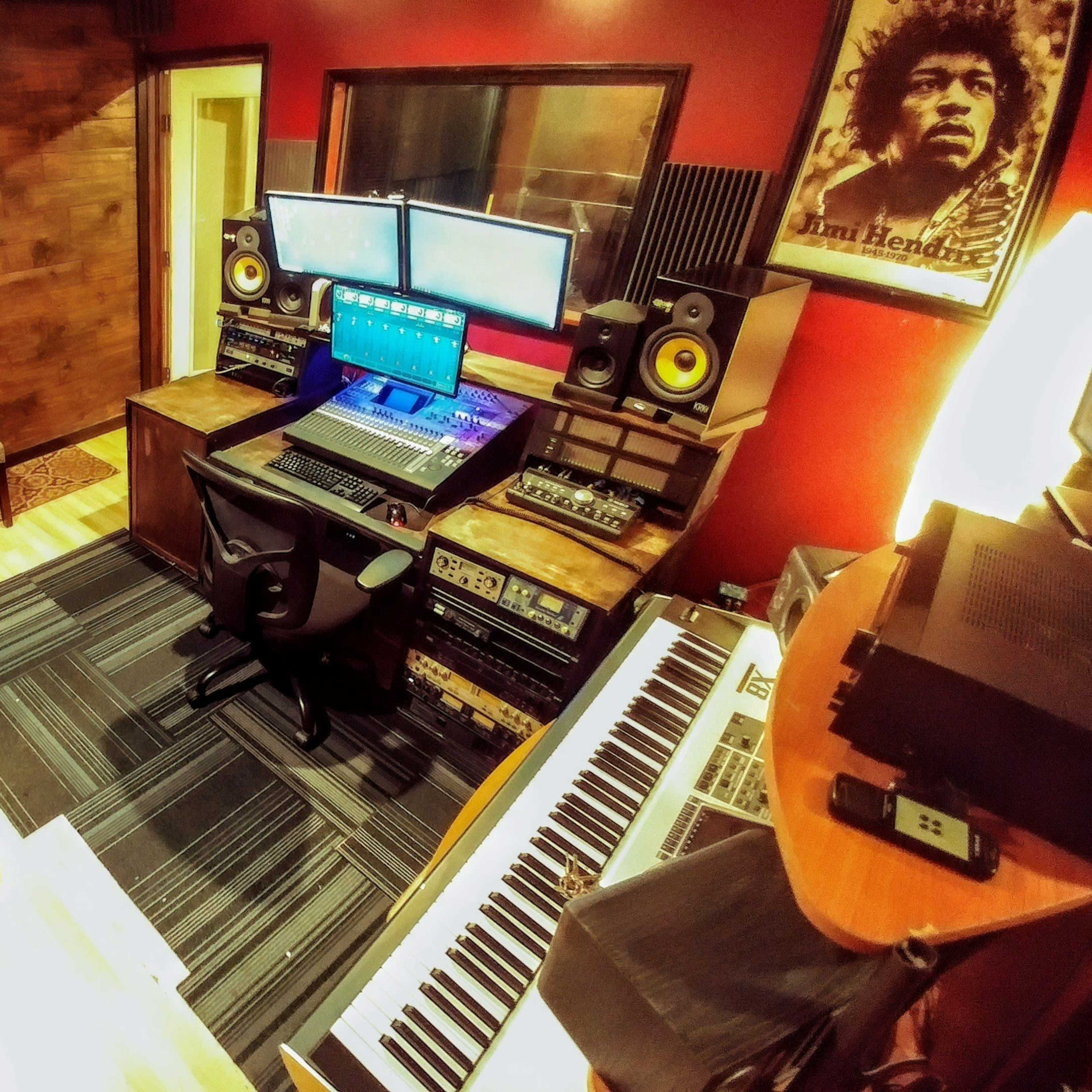 A music studio with a keyboard, dual computer monitors, mixing equipment, and several guitars displayed on the wall.