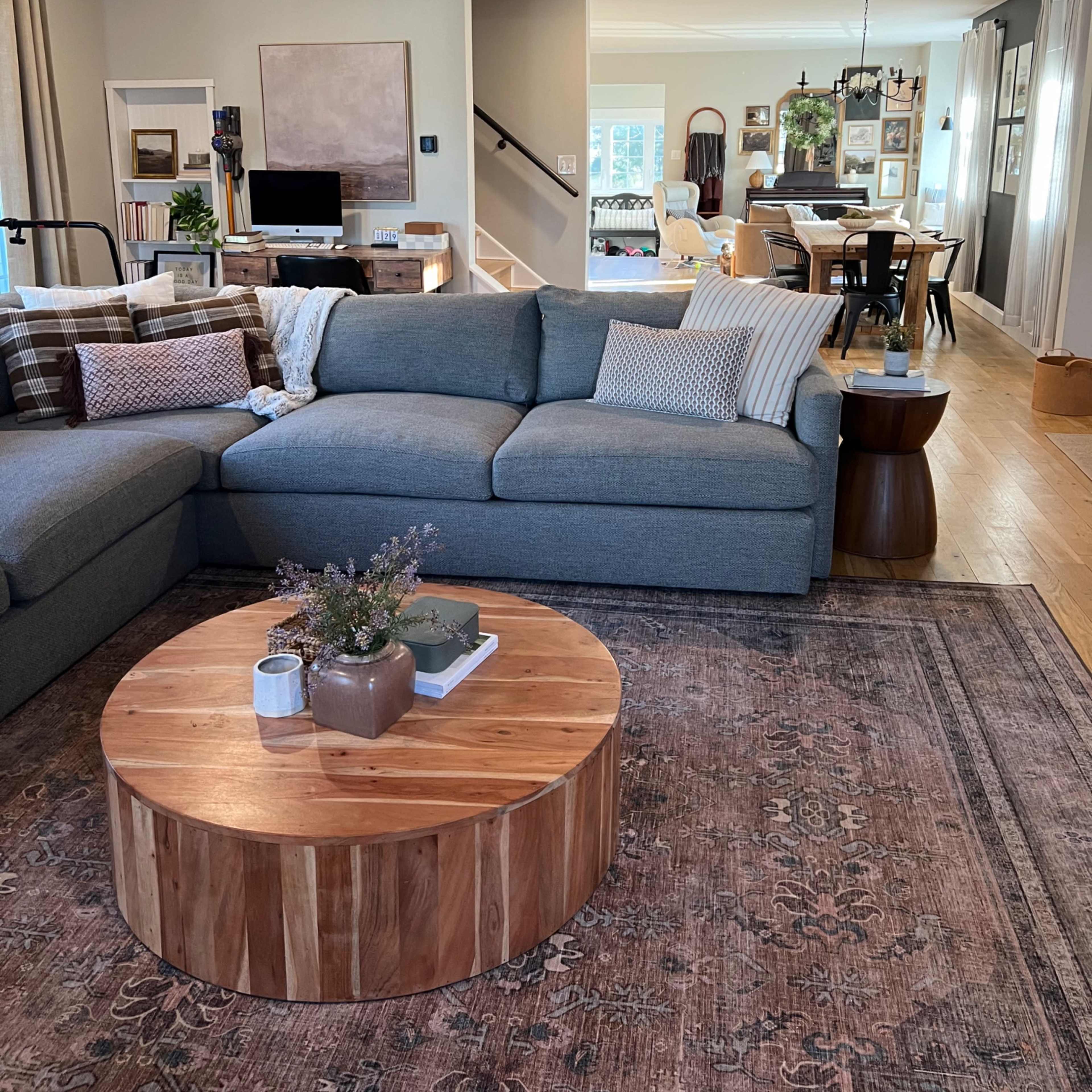A spacious living room features a grey sectional sofa, a circular wooden coffee table, and an intricate area rug, leading into a dining area visible in the background.