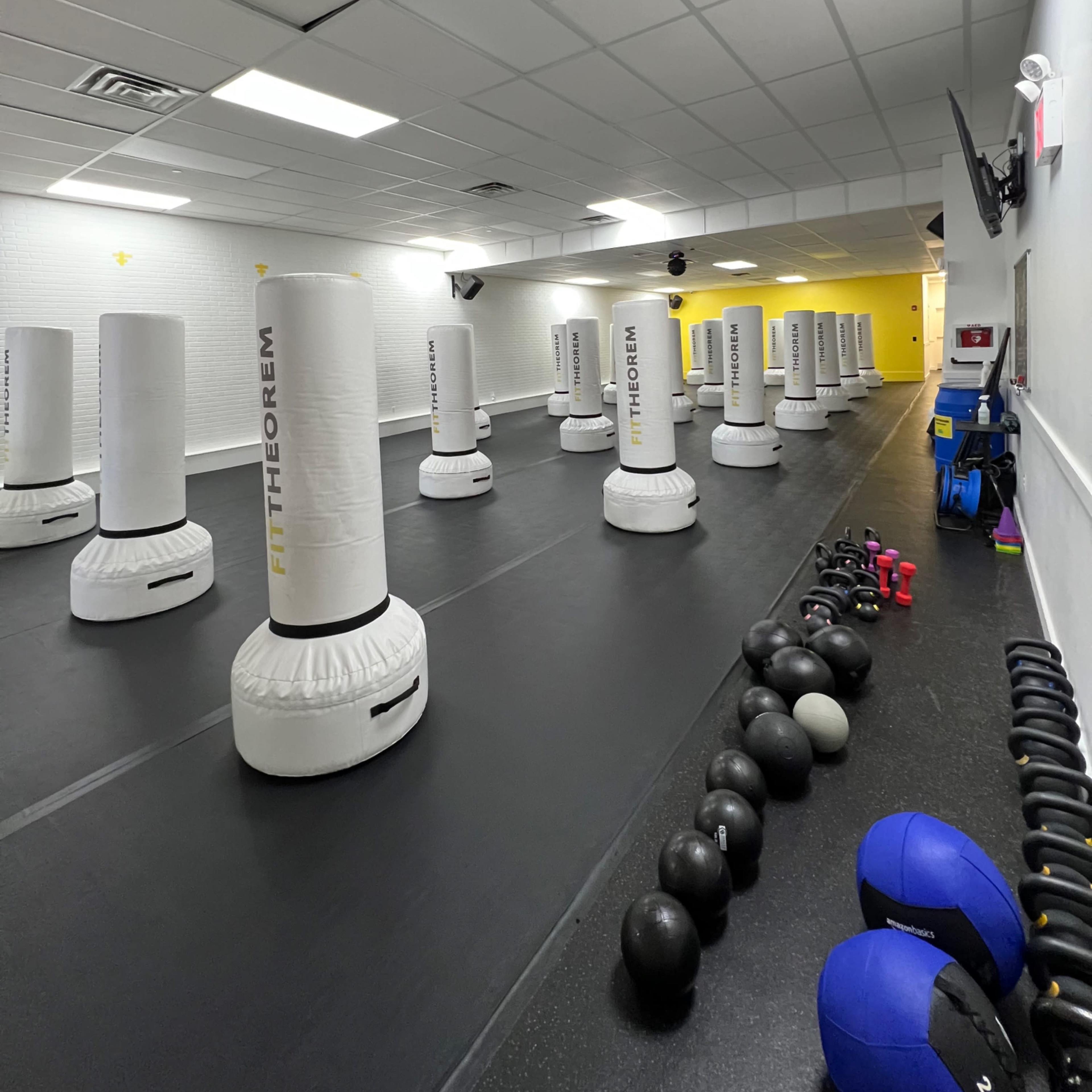 A fitness studio with multiple punching bags arranged on a black rubber floor, along with kettlebells and exercise balls lined against the wall.