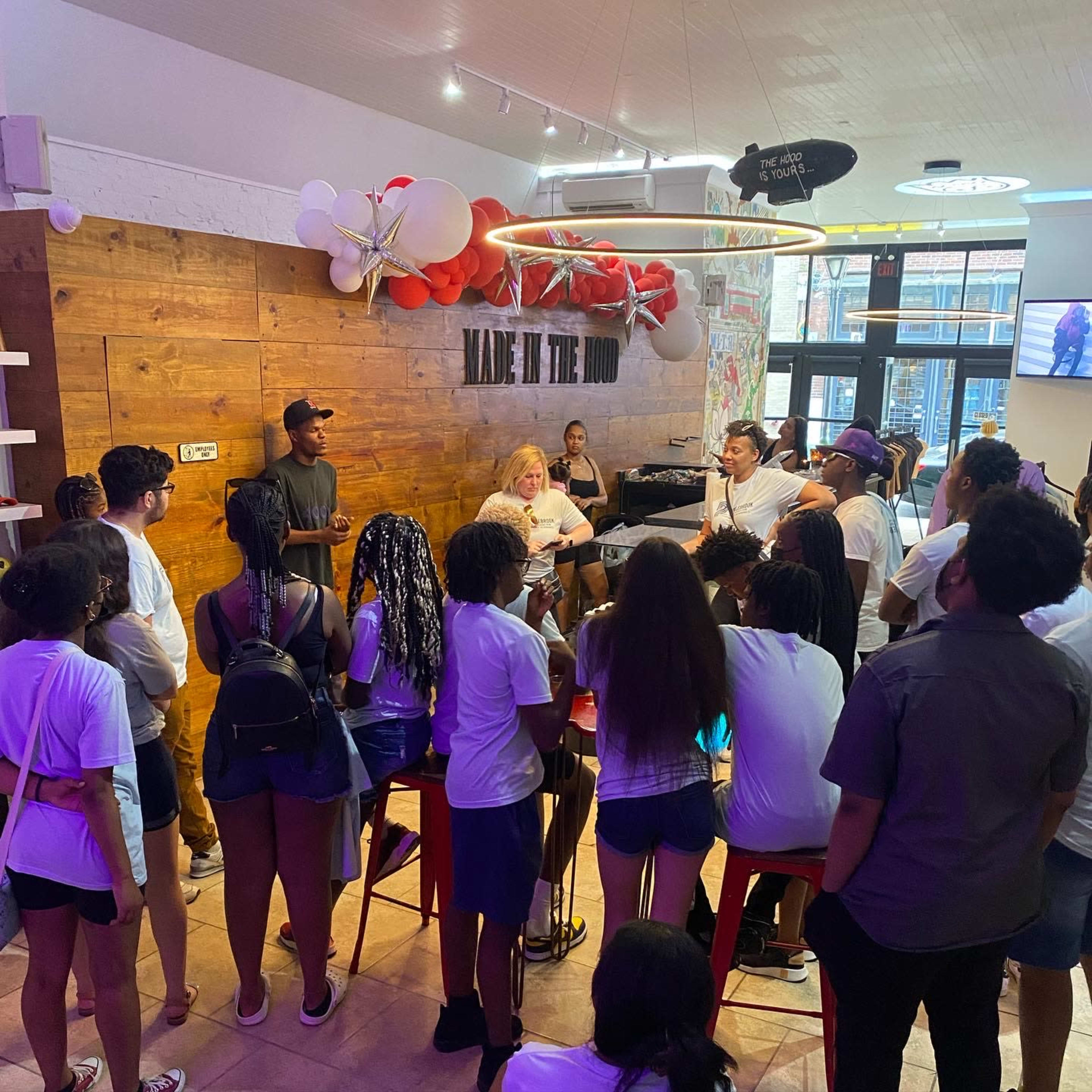 A group of people stands in front of a counter in a brightly lit café with wooden decor, listening to a speaker.