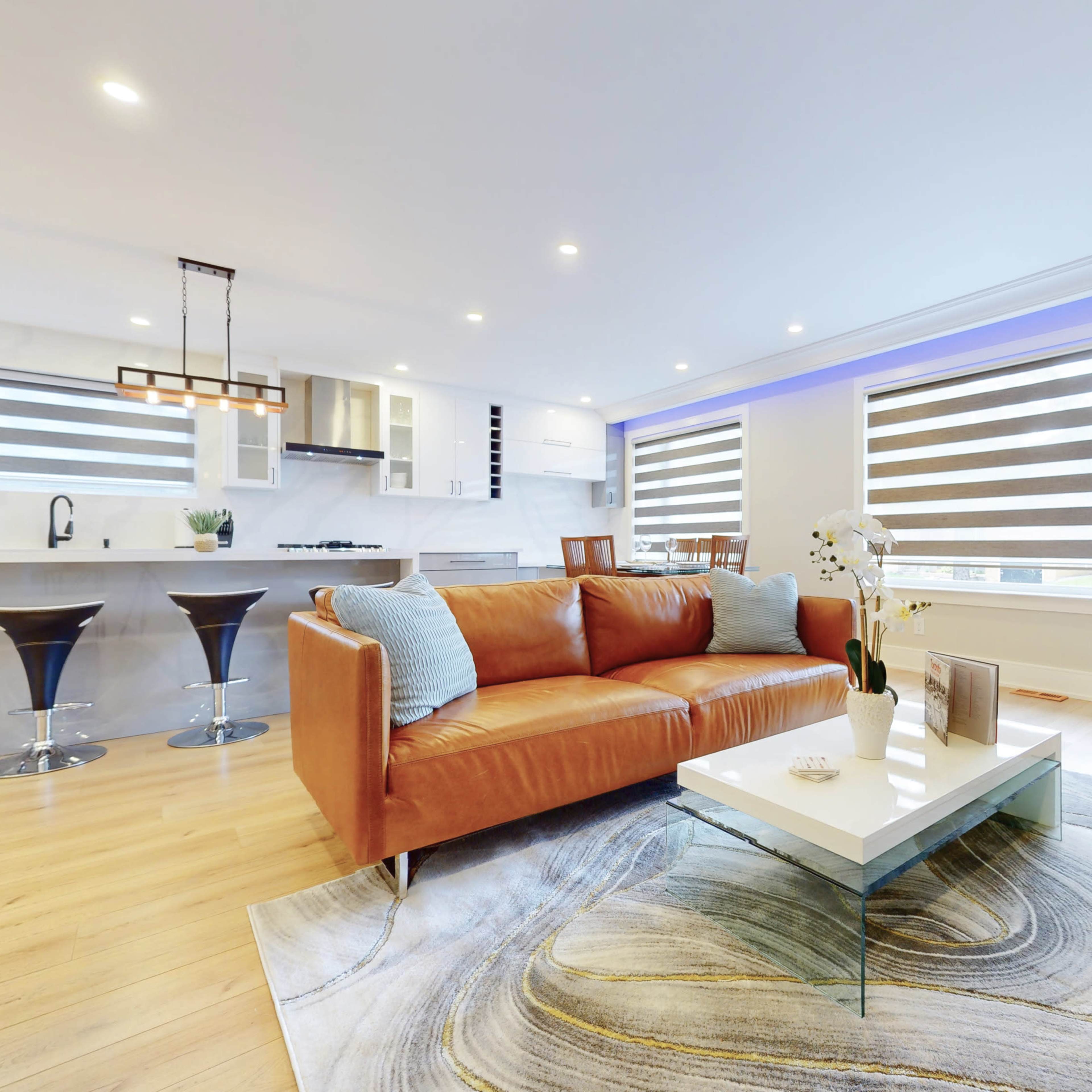 A modern living room featuring a brown leather sofa, a glass coffee table on a patterned rug, and a kitchen area in the background with sleek appliances.