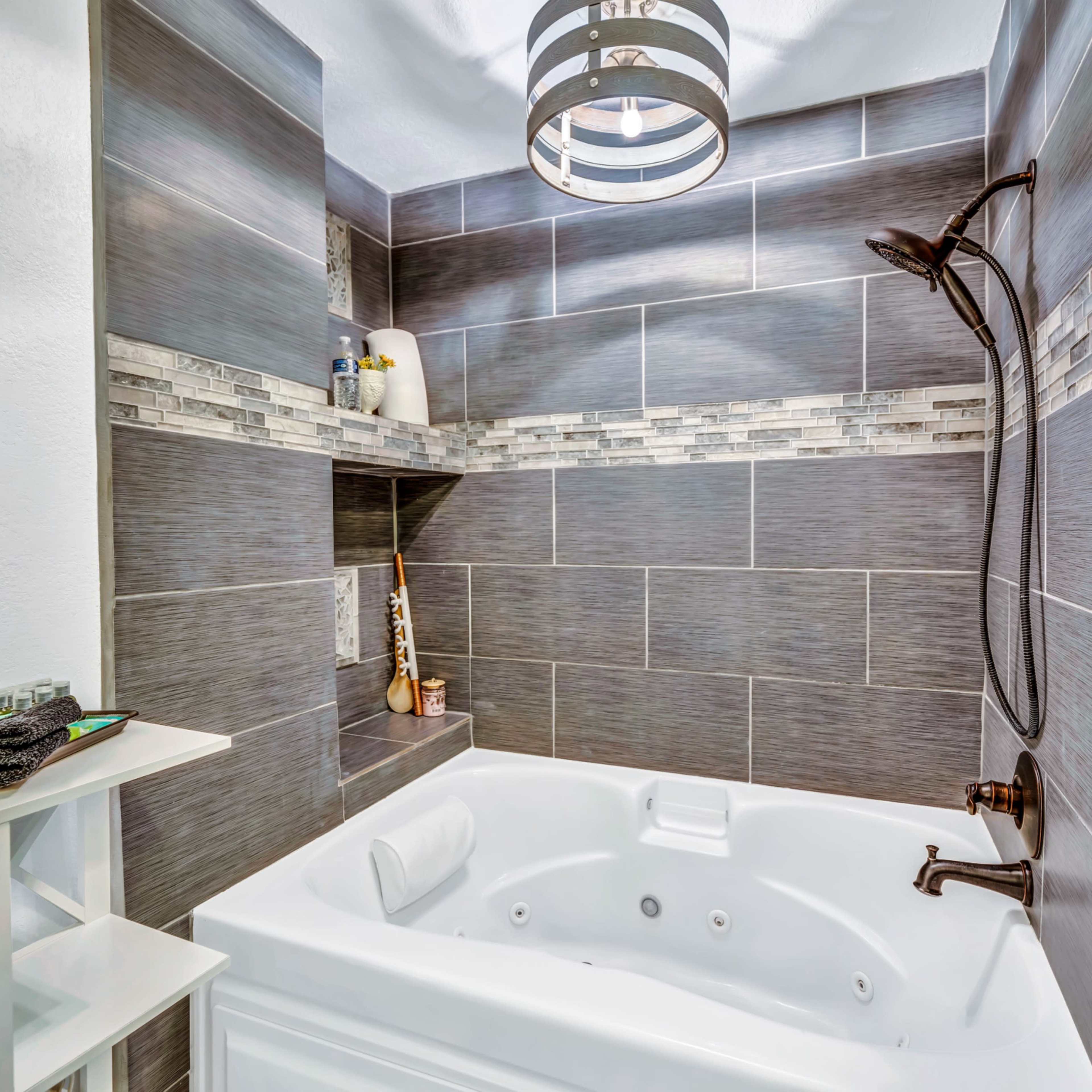 A white, jetted bathtub is positioned against a wall of gray tiled panels, featuring a decorative tile border and a modern light fixture overhead.