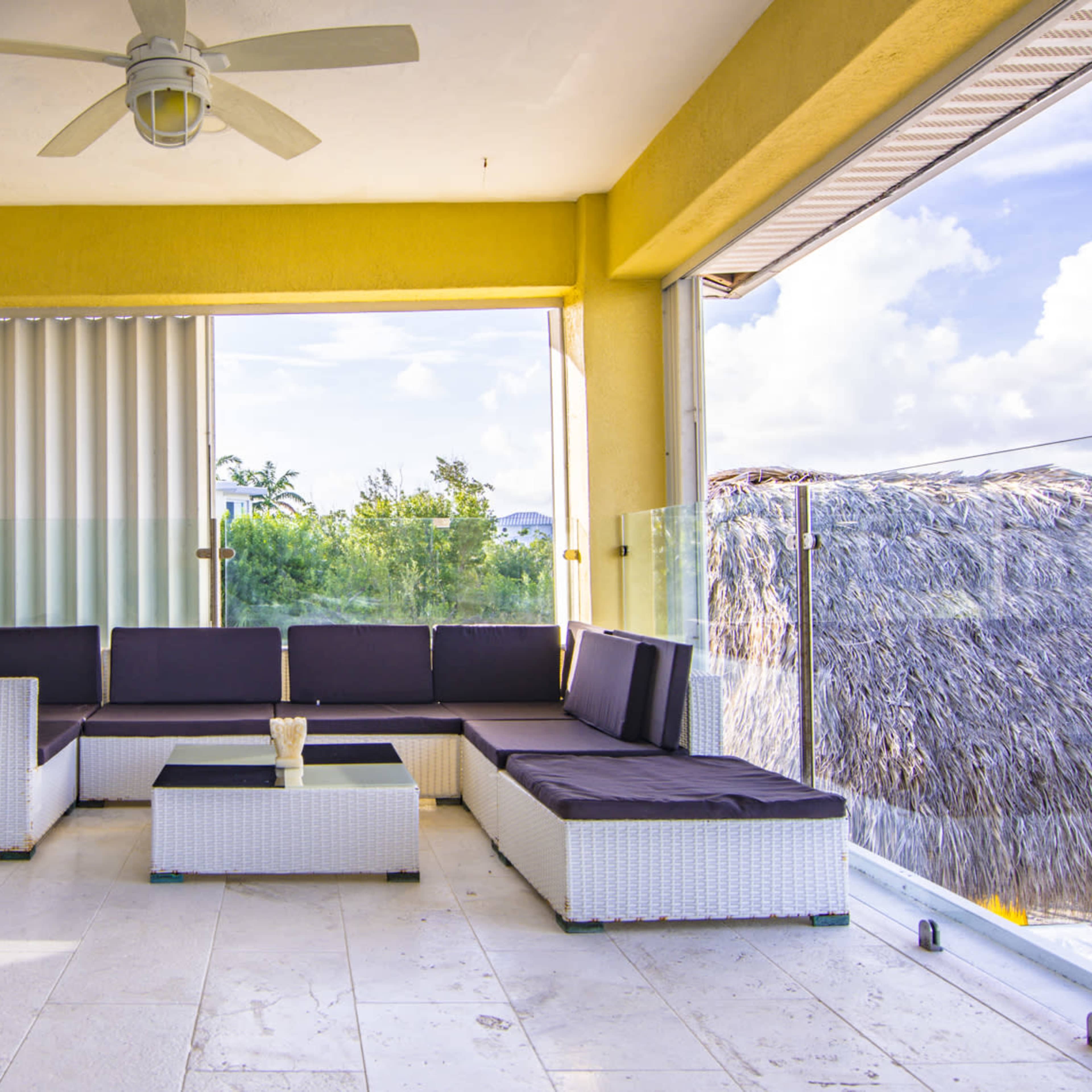 A modern, covered outdoor seating area with a sectional sofa and a central table, featuring large sliding glass doors and a view of greenery outside.