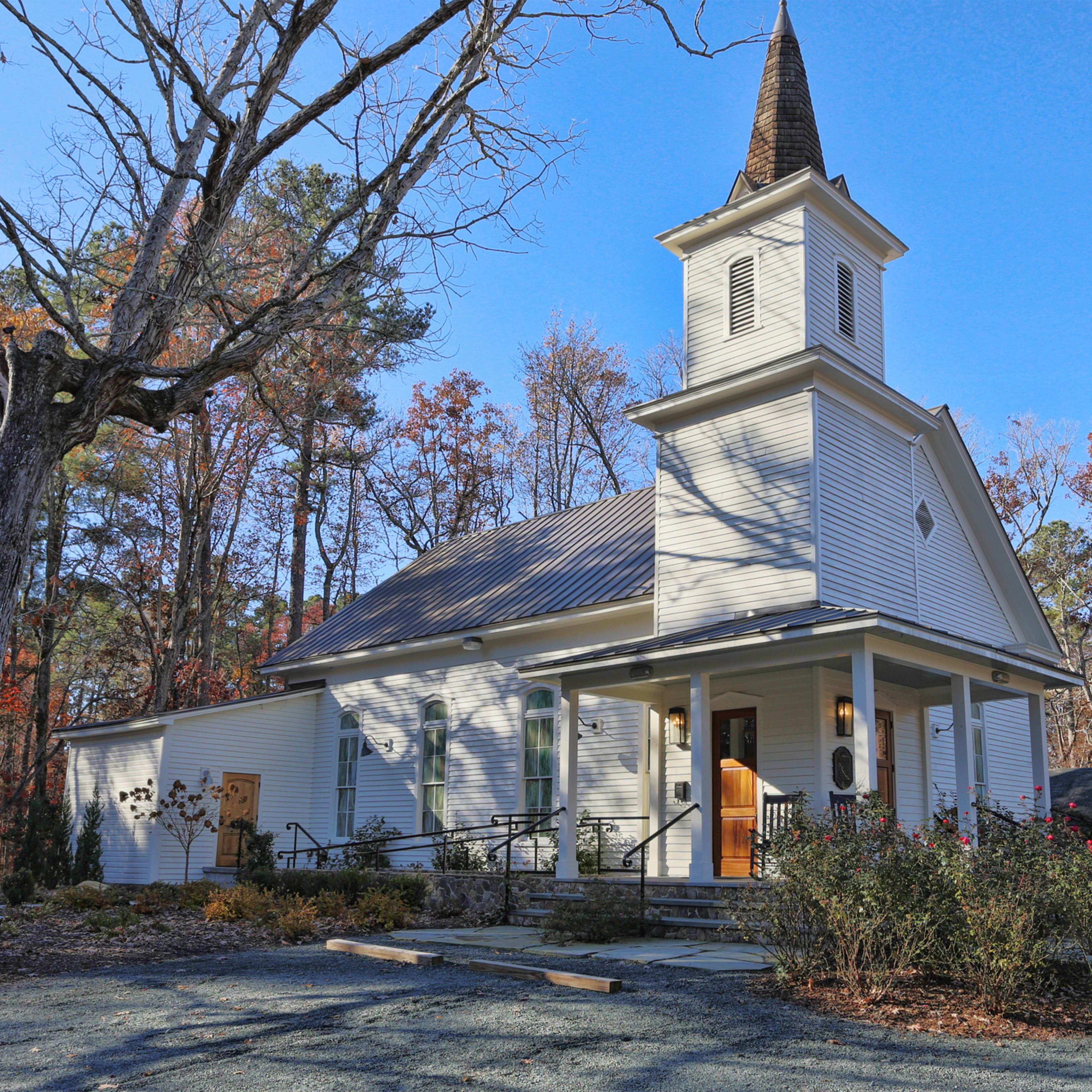 A white church with a tall spire, surrounded by trees and a gravel walkway.