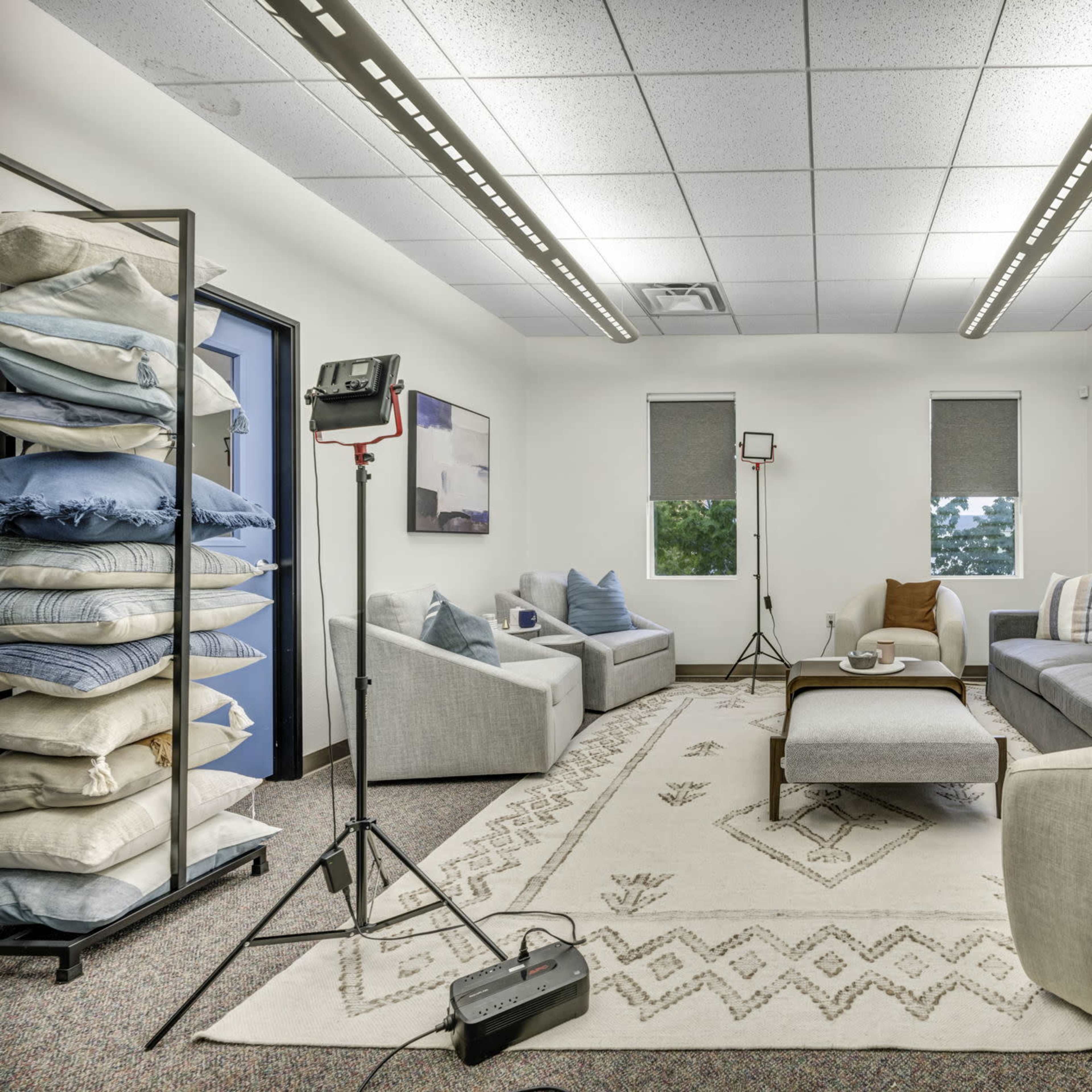 A well-lit room features two couches facing each other, a coffee table in the center, and a stack of pillows against one wall, with lighting equipment set up for a photo shoot.