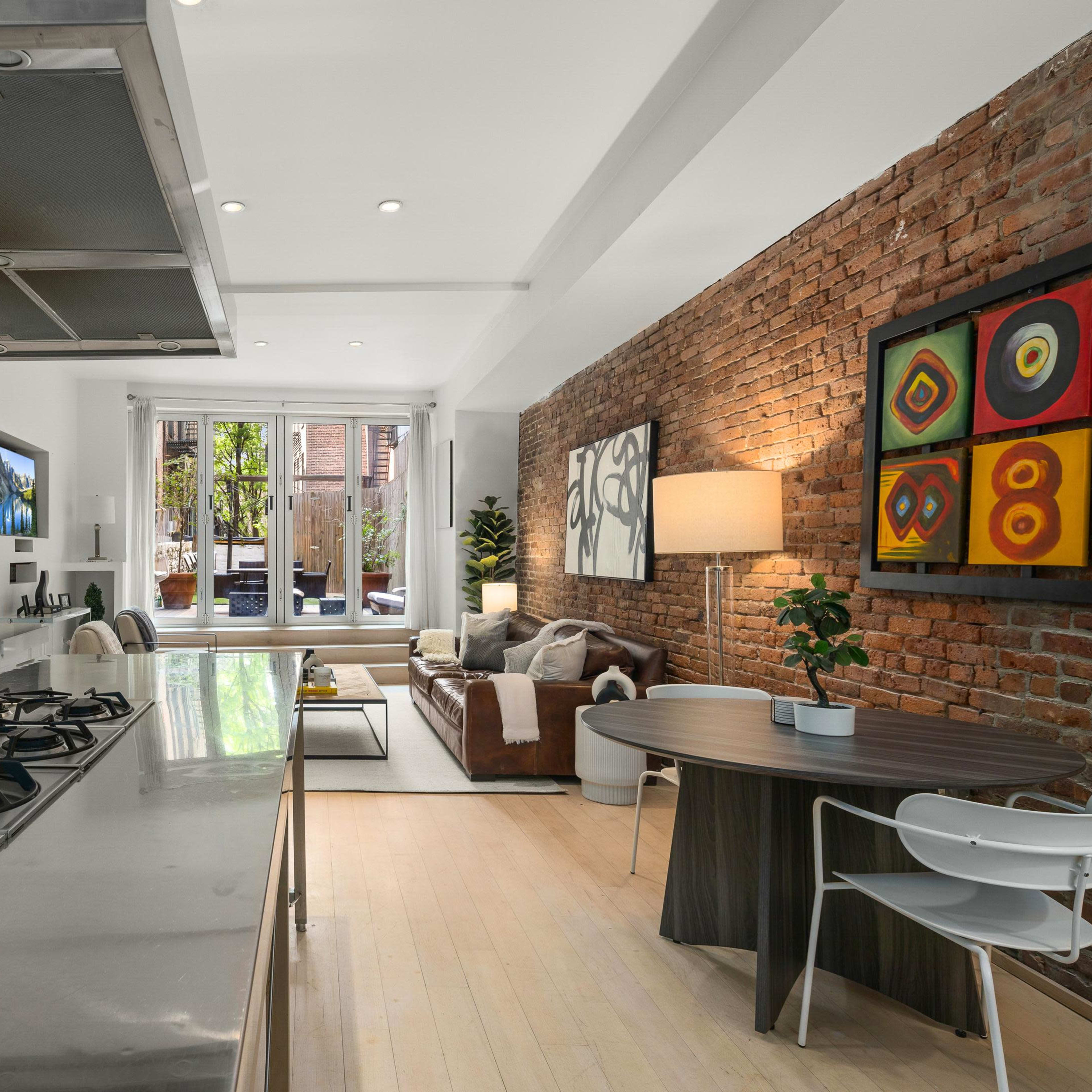 A modern kitchen with a stainless-steel countertop and gas stove, adjacent to a living area featuring a brown sofa and a view of an outdoor space through large windows.