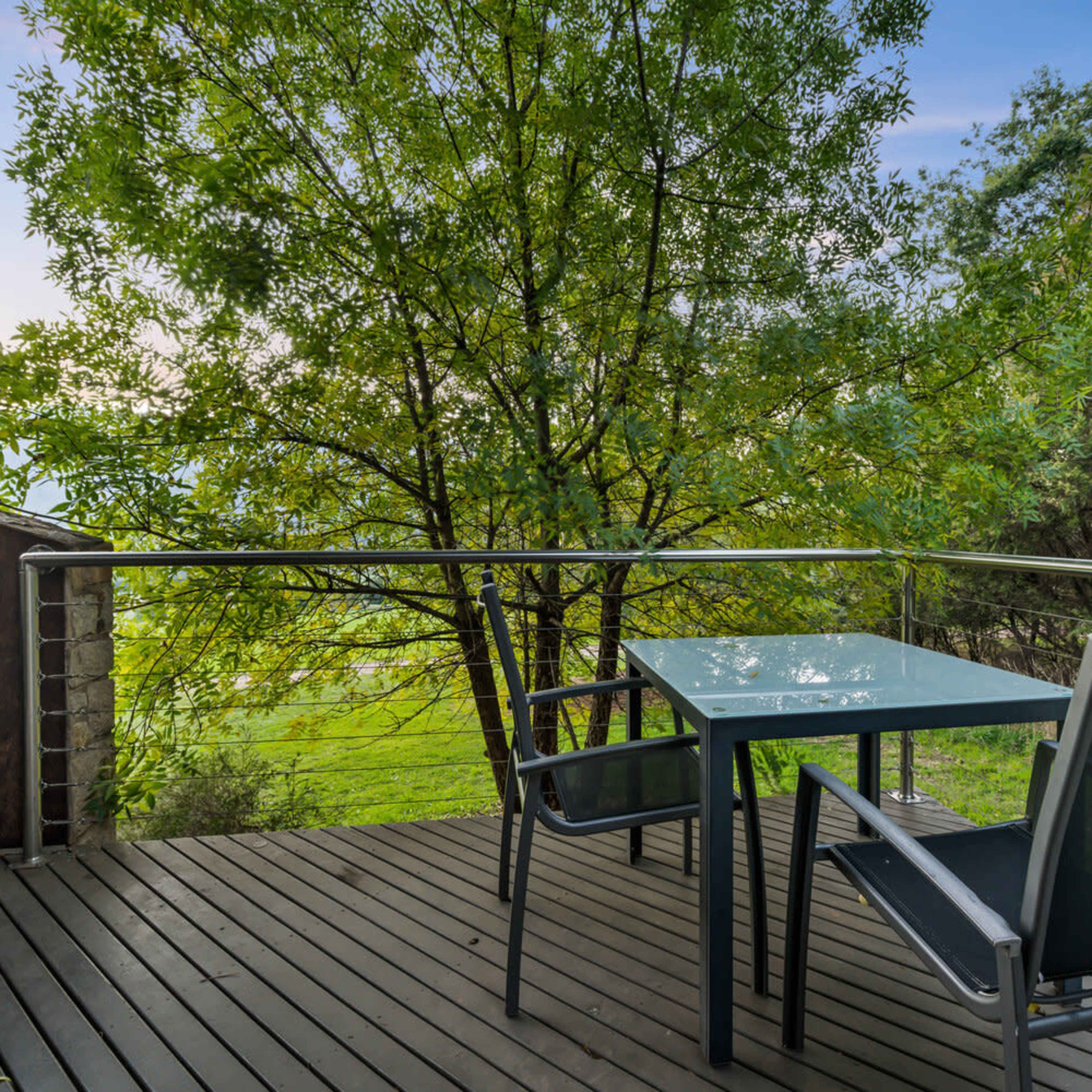 A metal table and two chairs are positioned on a wooden deck surrounded by green foliage and an expansive view of grass and hills.