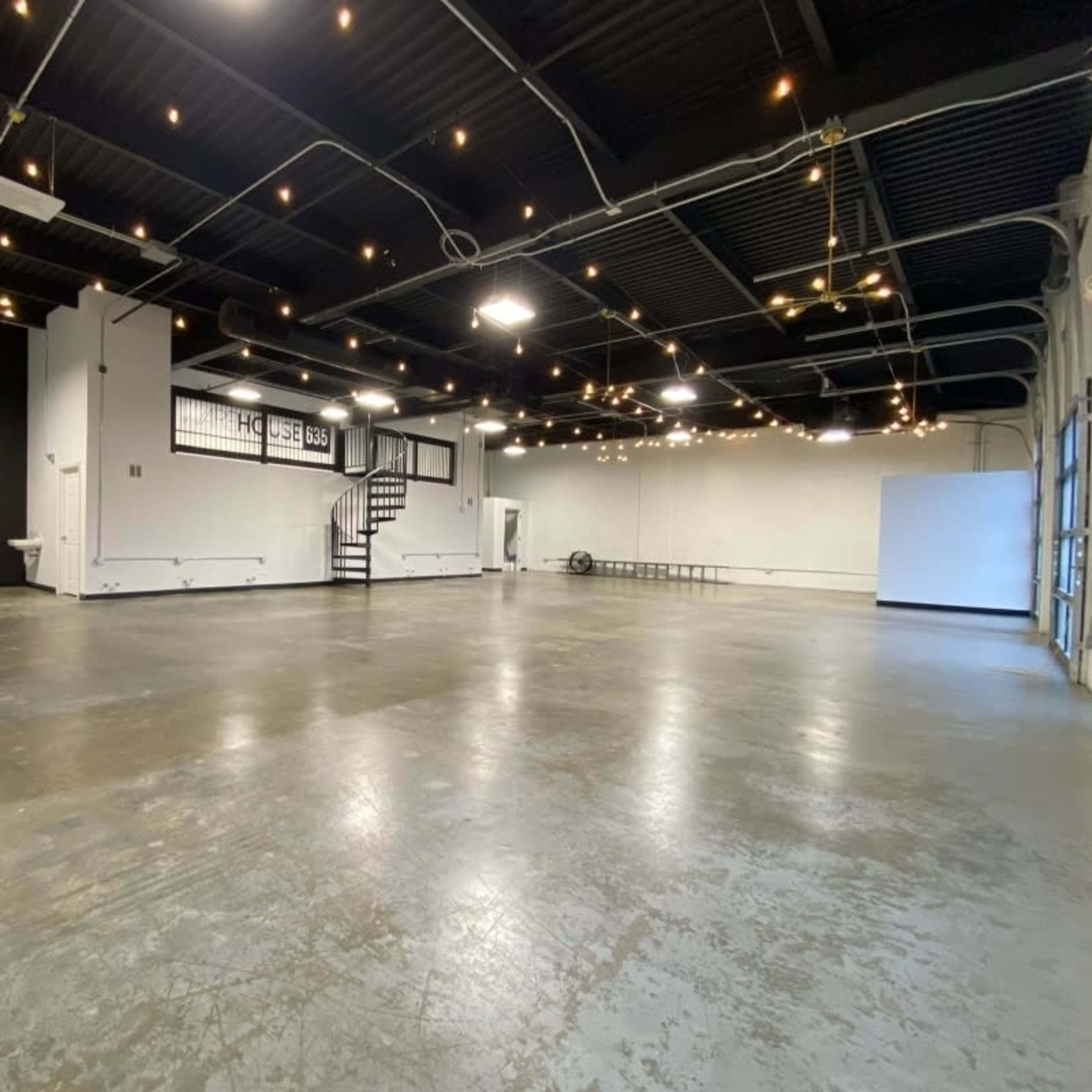 A spacious, empty room with concrete flooring, exposed black ceiling, and a staircase in the background, illuminated by string lights.