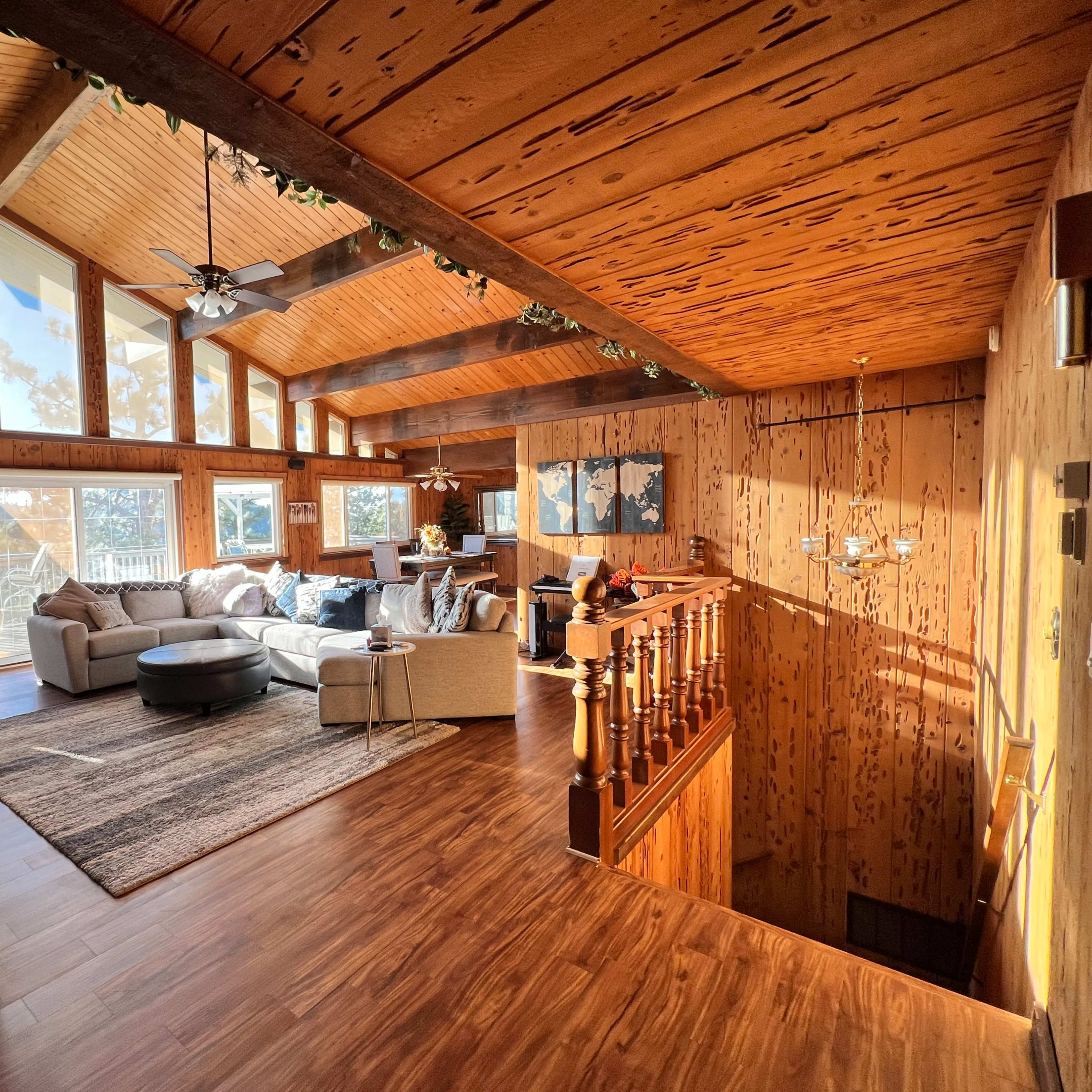 A spacious living room with wooden walls and ceiling, large windows allowing natural light, and a cozy seating area featuring a sectional sofa and coffee table.