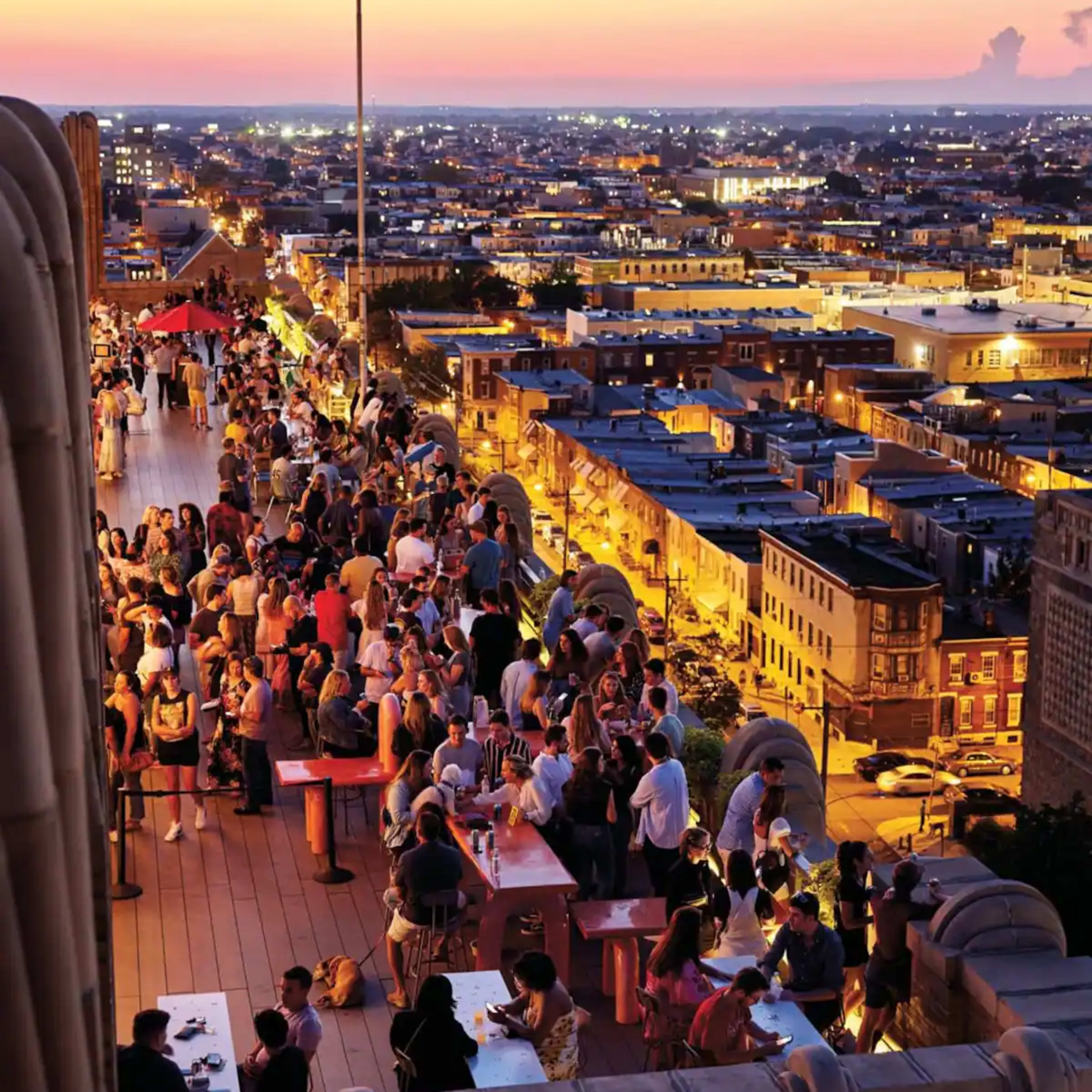 A large crowd gathers on a rooftop terrace overlooking a city skyline illuminated by twilight.