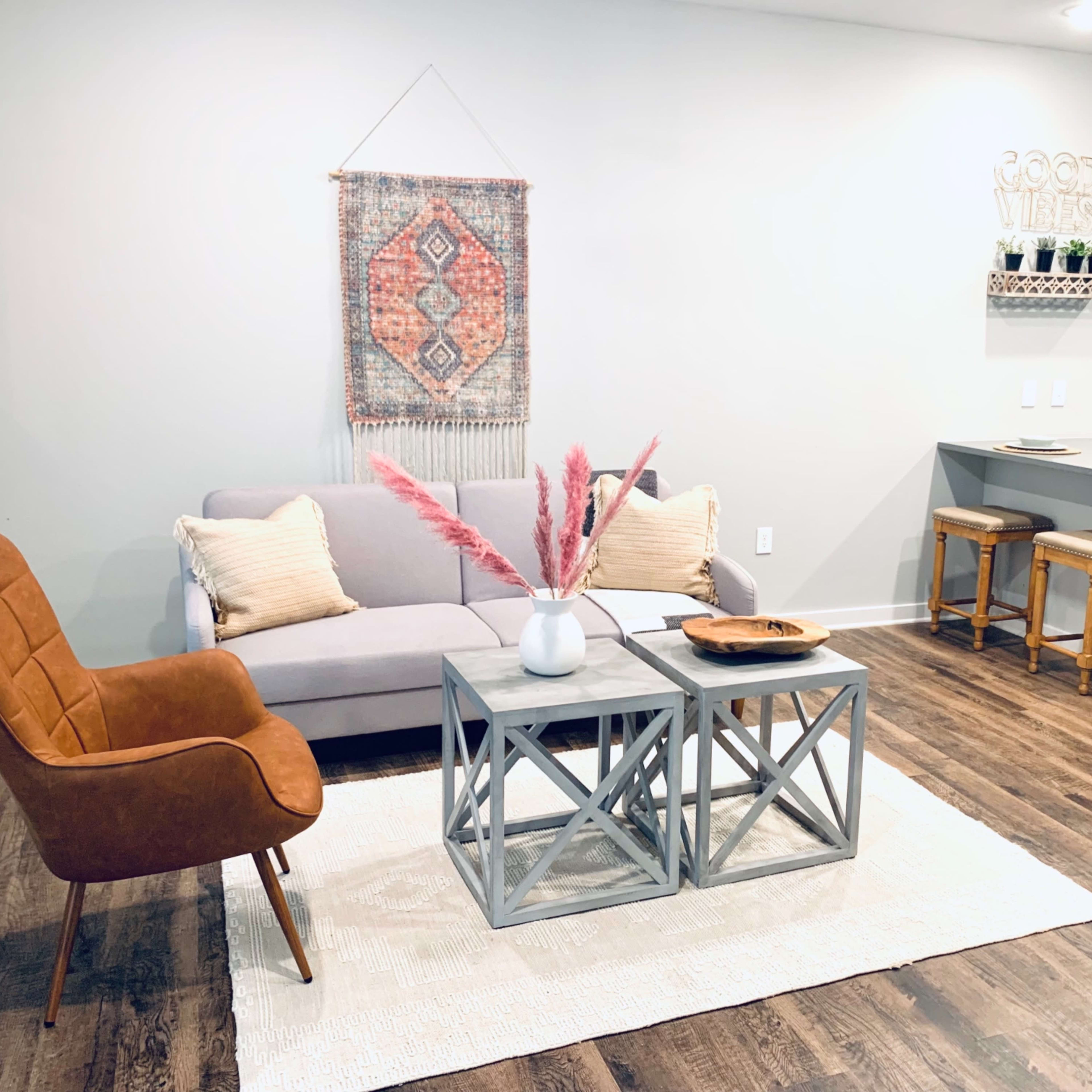 A modern living room with a gray sofa, a wooden coffee table, a decorative wall hanging, and a kitchen bar in the background.
