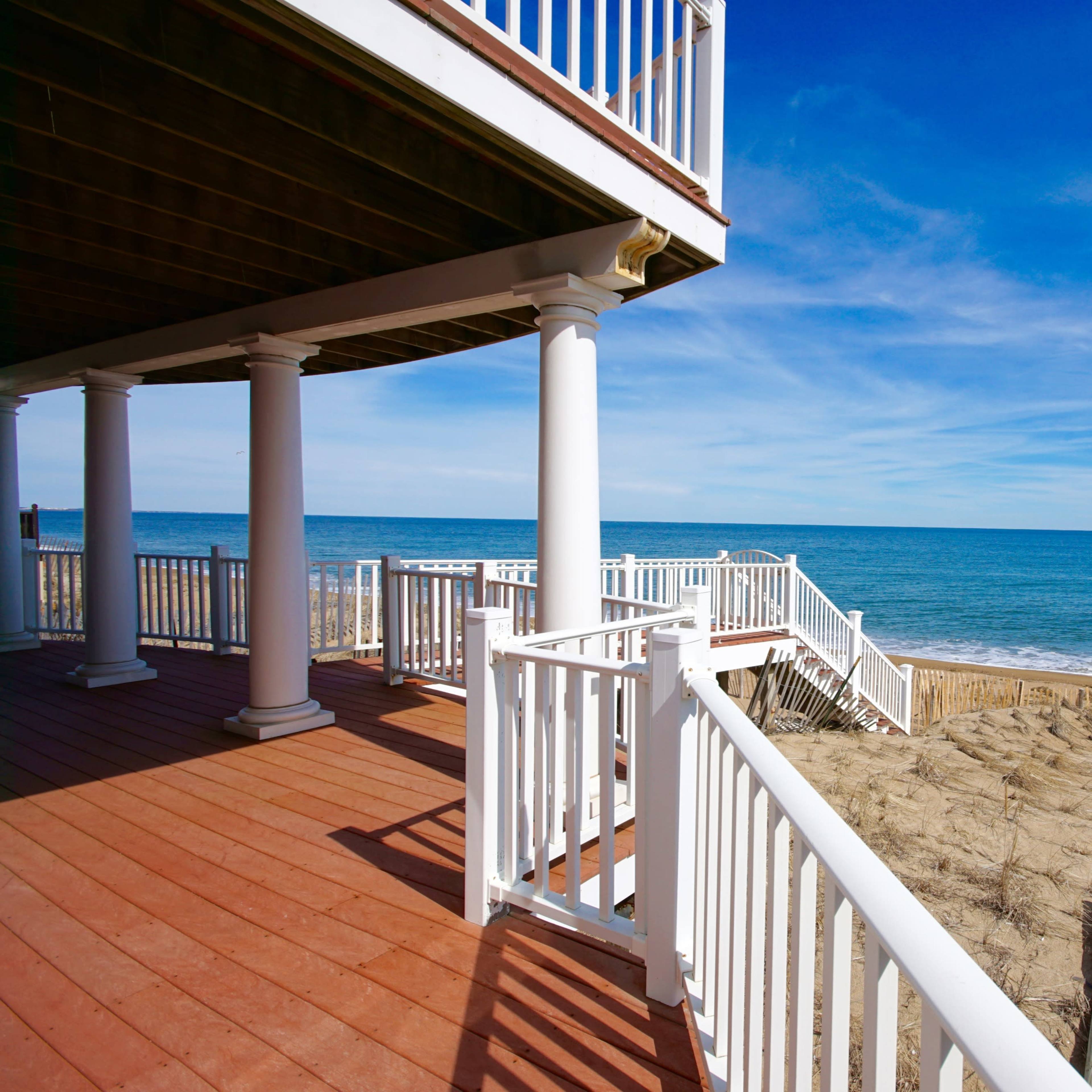 A wooden deck with white railings overlooks a sandy beach and the ocean under a clear blue sky.