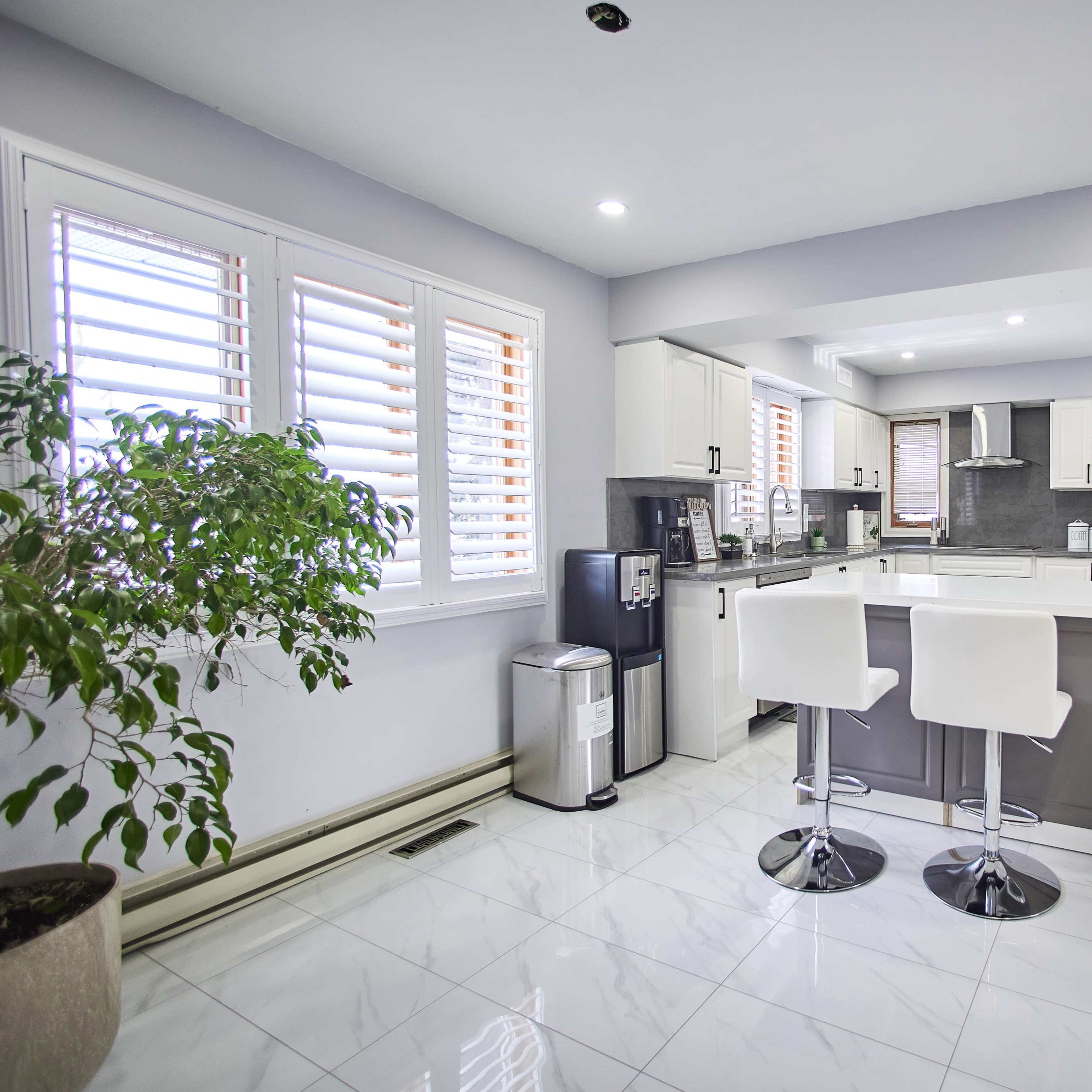 A modern kitchen featuring white cabinets, a gray island with bar stools, and large windows with shutter blinds.