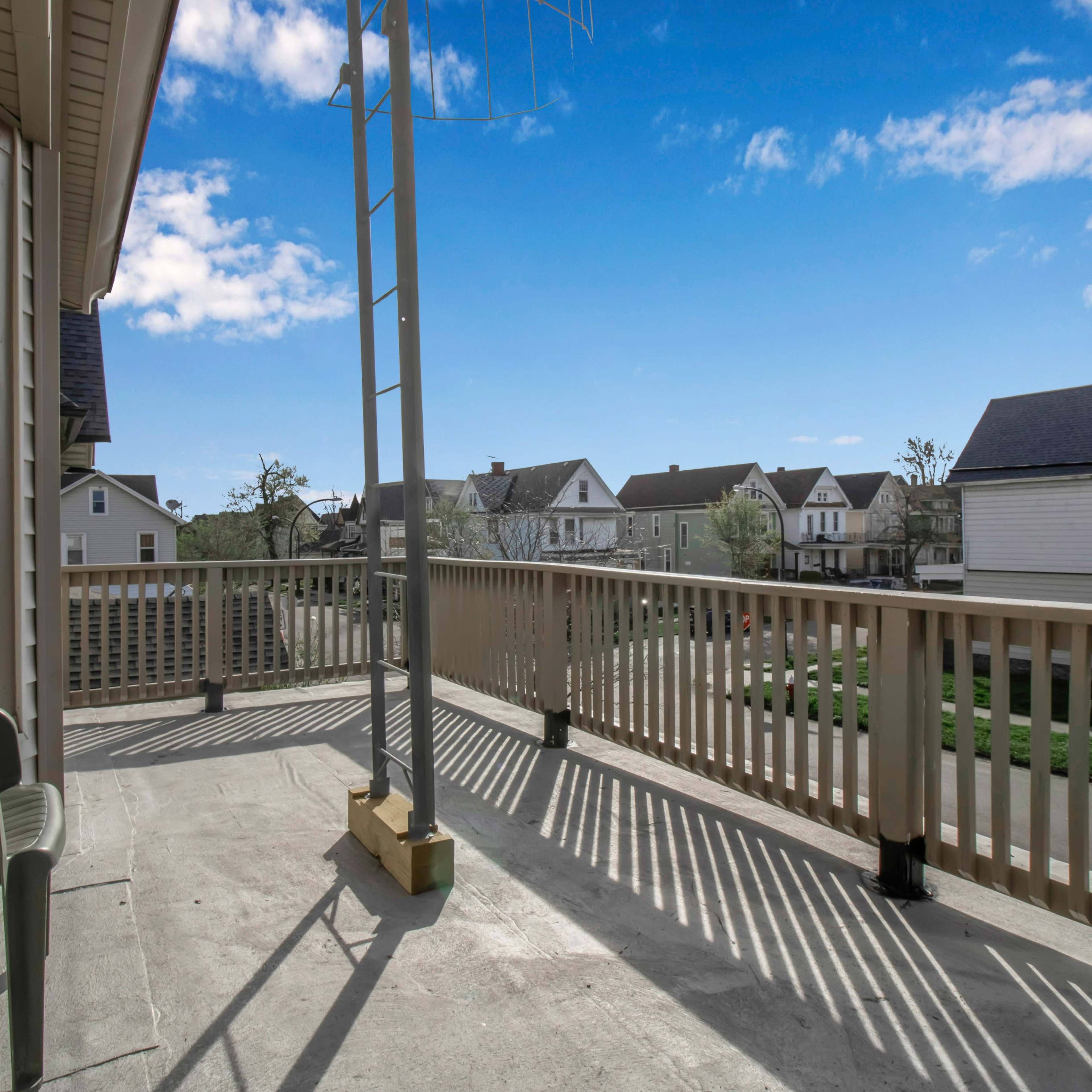 A balcony with a green chair overlooks a neighborhood of houses under a blue sky with scattered clouds.