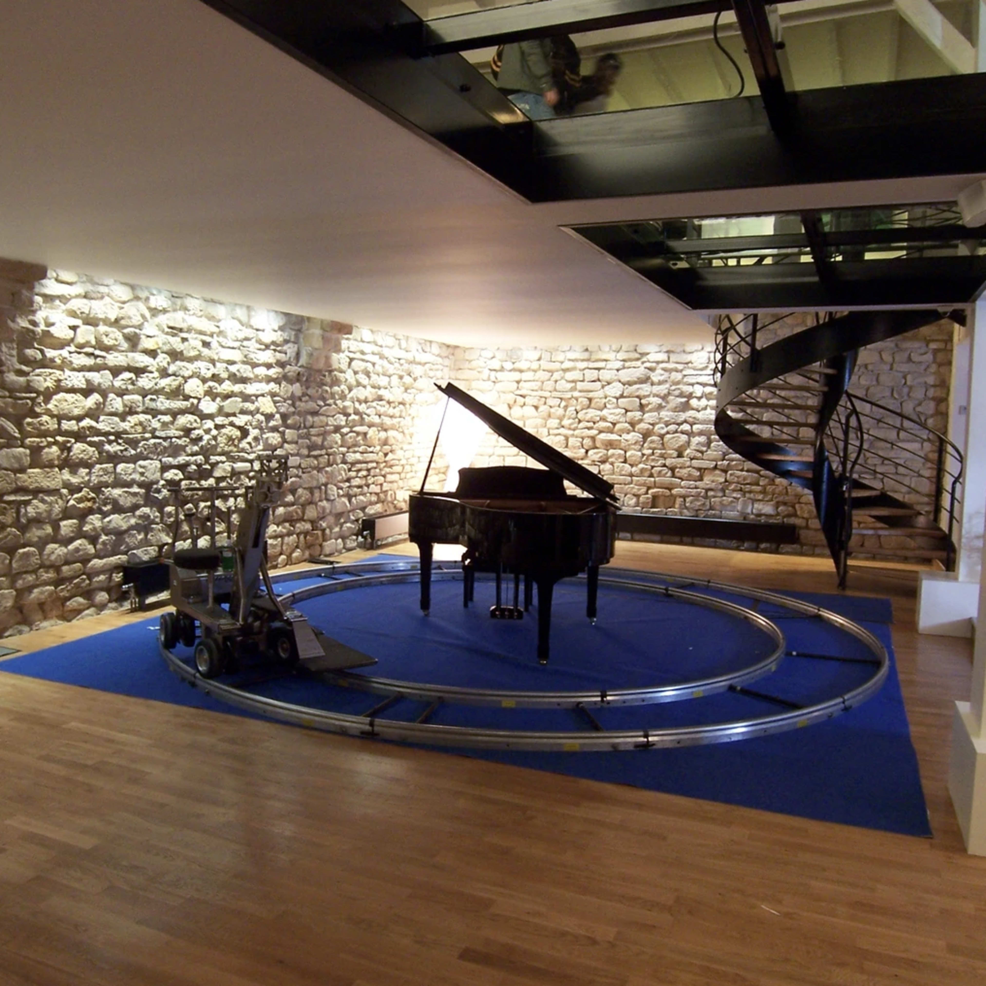 A grand piano situated on a circular platform with a track, surrounded by a wooden floor and stone walls in a spacious interior.