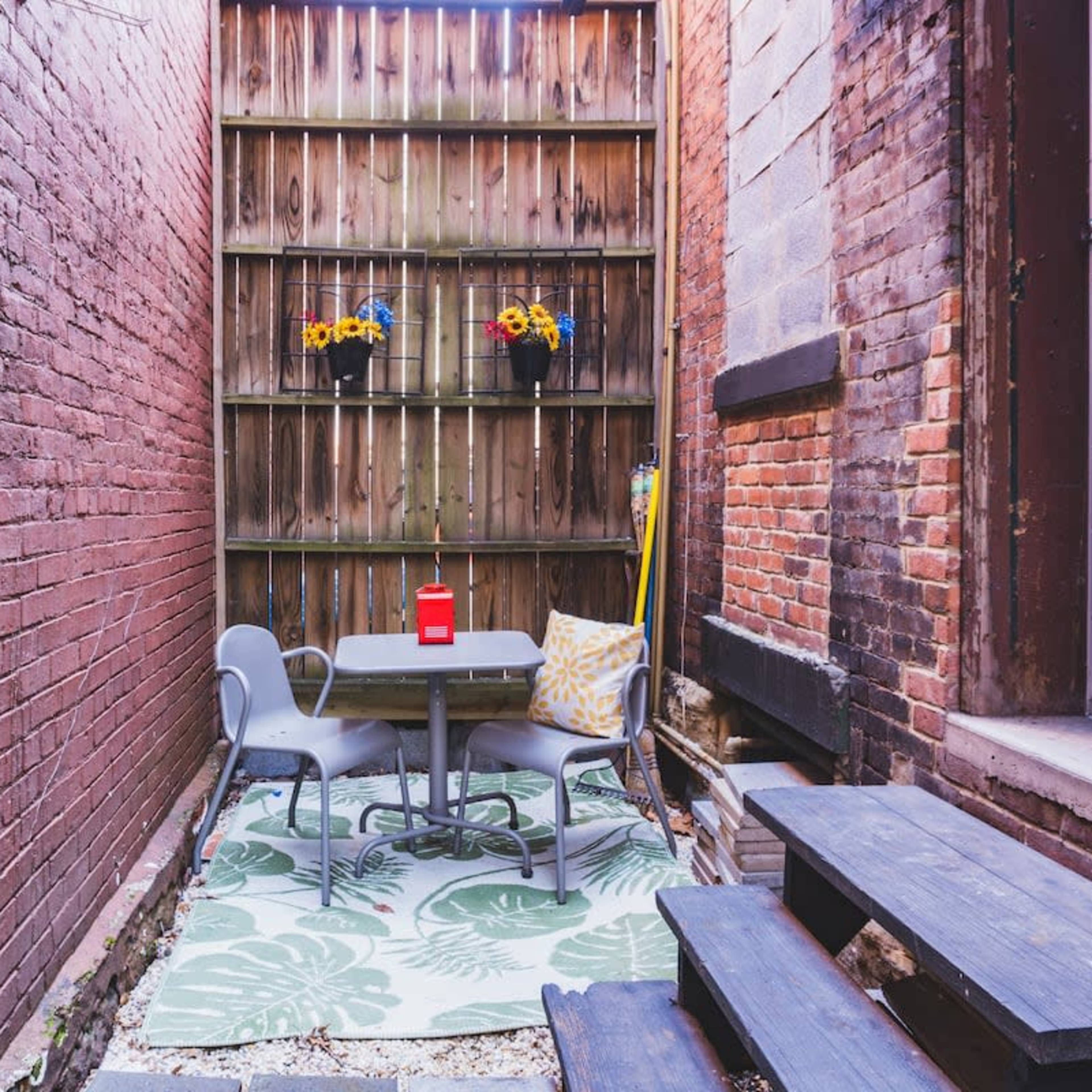 A small outdoor seating area is enclosed by brick walls, featuring a table and two chairs on a patterned rug, with flower pots hanging on the wooden fence above.