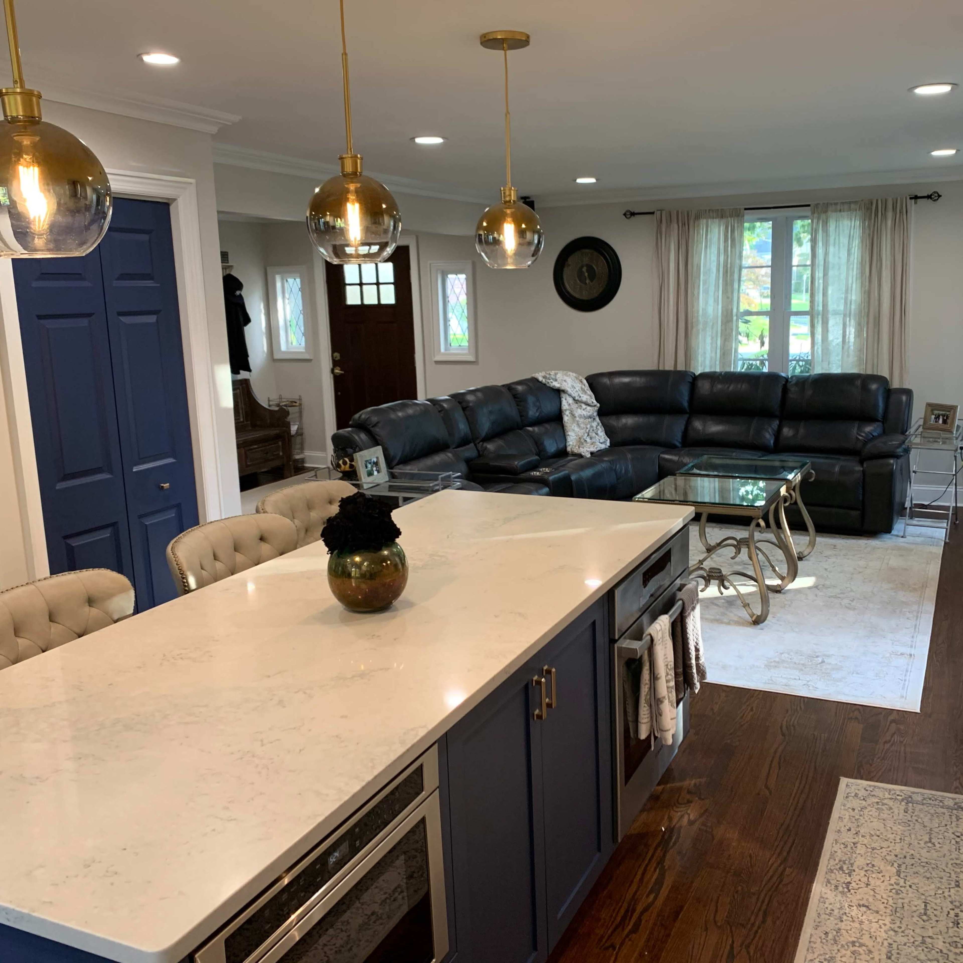 A modern kitchen with a marble countertop island, a black leather sectional sofa, and pendant lights illuminating the space.