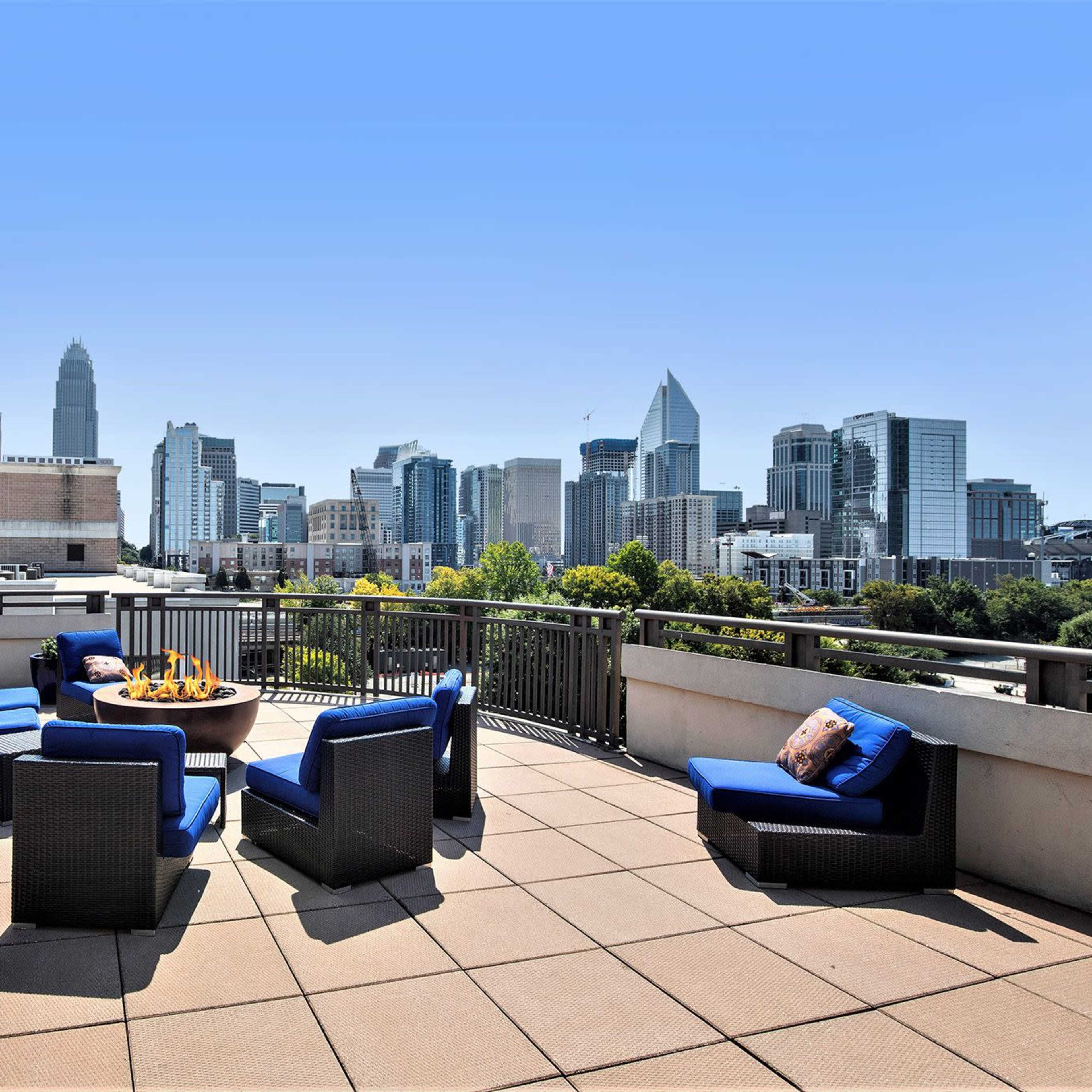 A rooftop terrace features seating around a fire pit with a view of a city skyline under a clear blue sky.