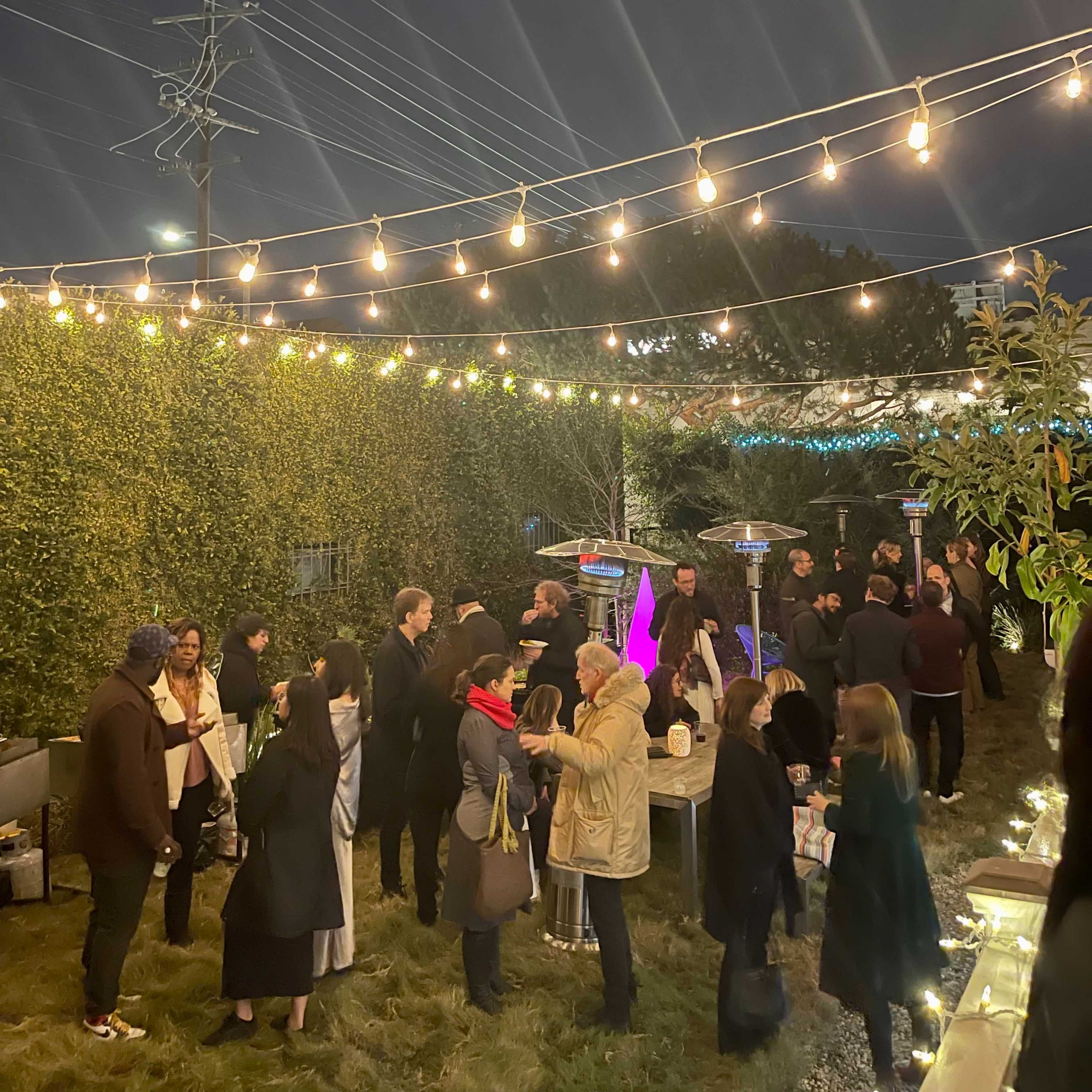 A crowd of people mingles under string lights in a garden setting at night, with a visible moon in the sky.