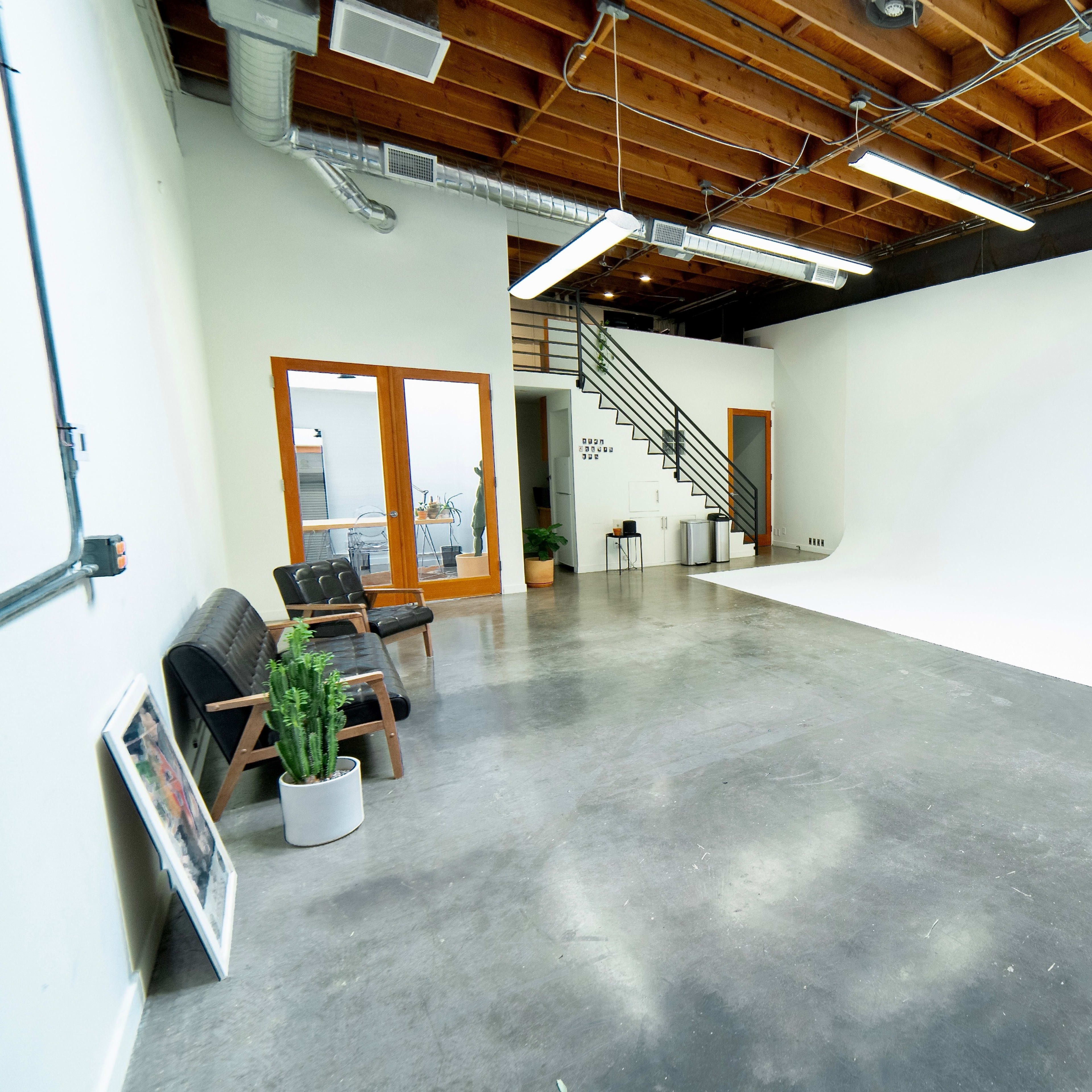 A spacious, empty studio with a white backdrop, concrete flooring, and a staircase leading to a second level.