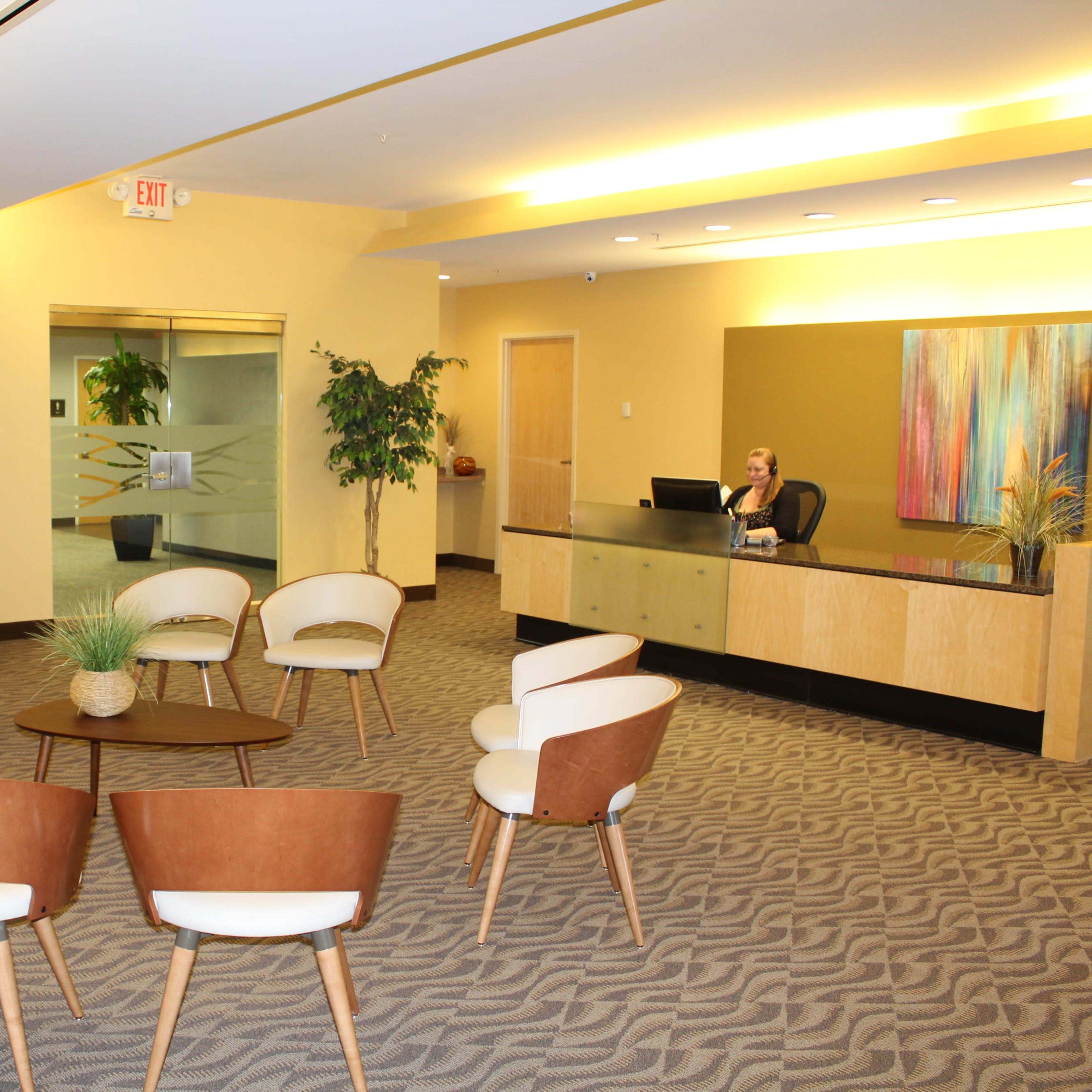 A modern reception area features a front desk with a seated receptionist, surrounded by several circular seating arrangements and a small coffee table.
