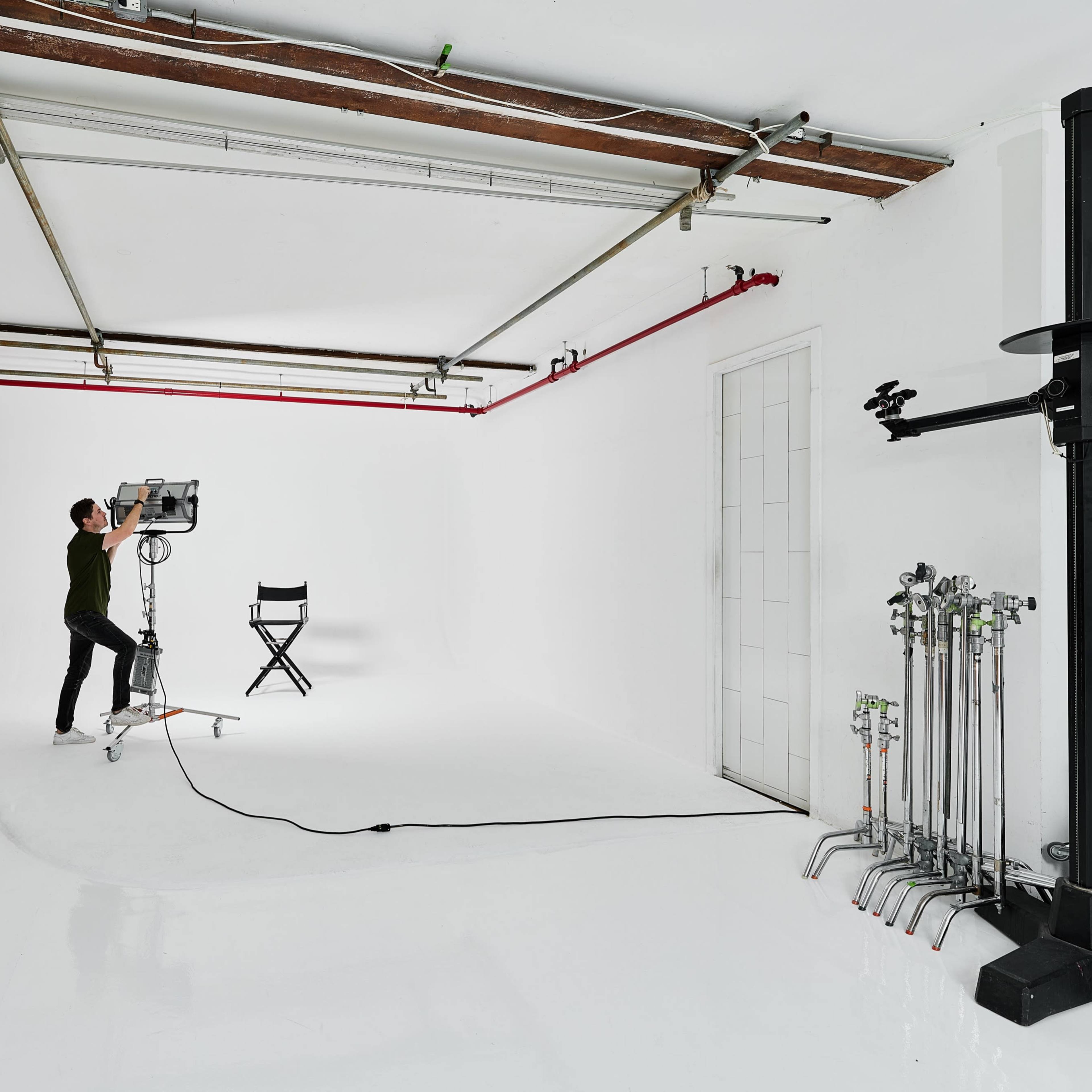 A person adjusts a lighting fixture in a blank studio space with a director's chair and various equipment along the side.