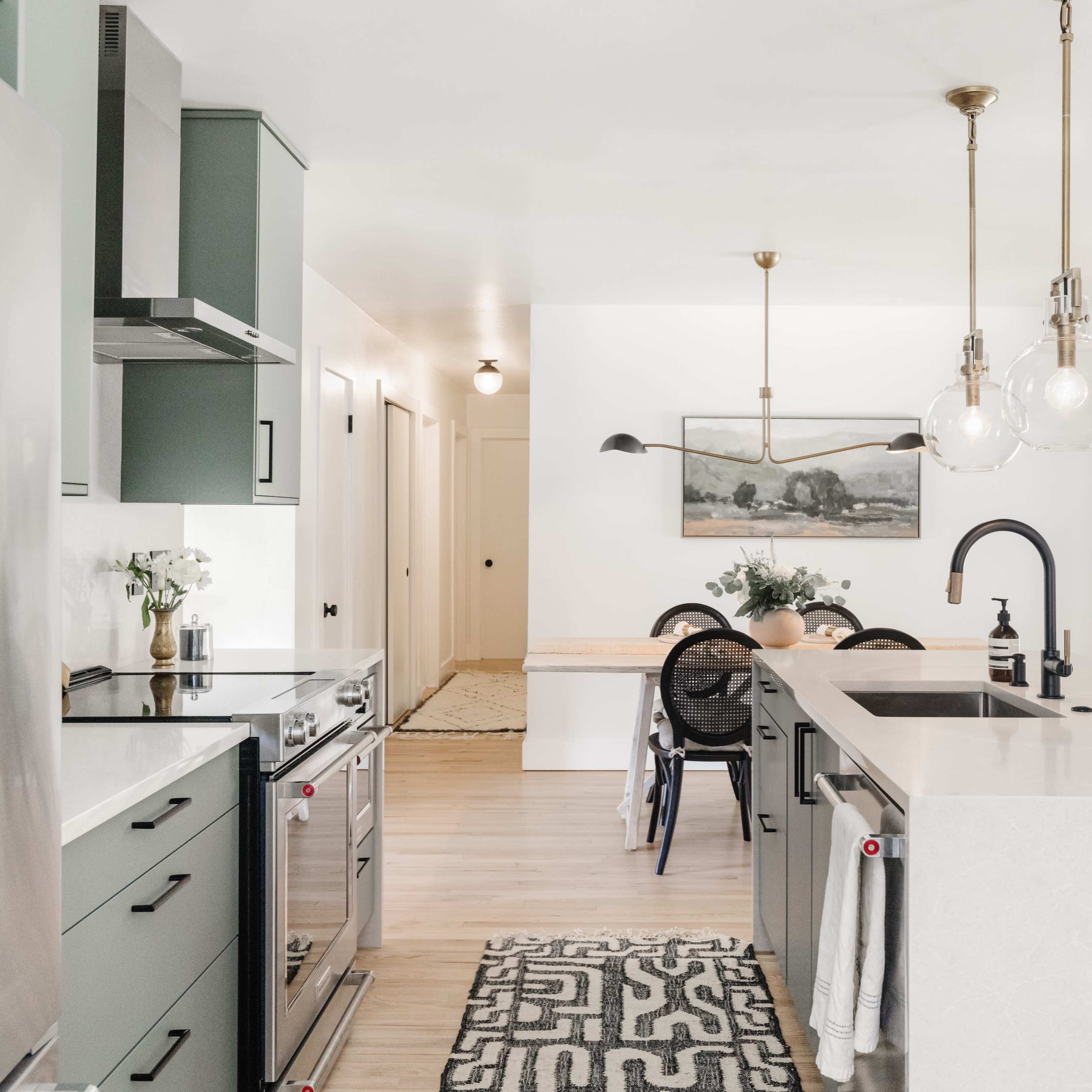A modern kitchen features green cabinets, stainless steel appliances, and a dining area with a wooden table and pendant lighting.